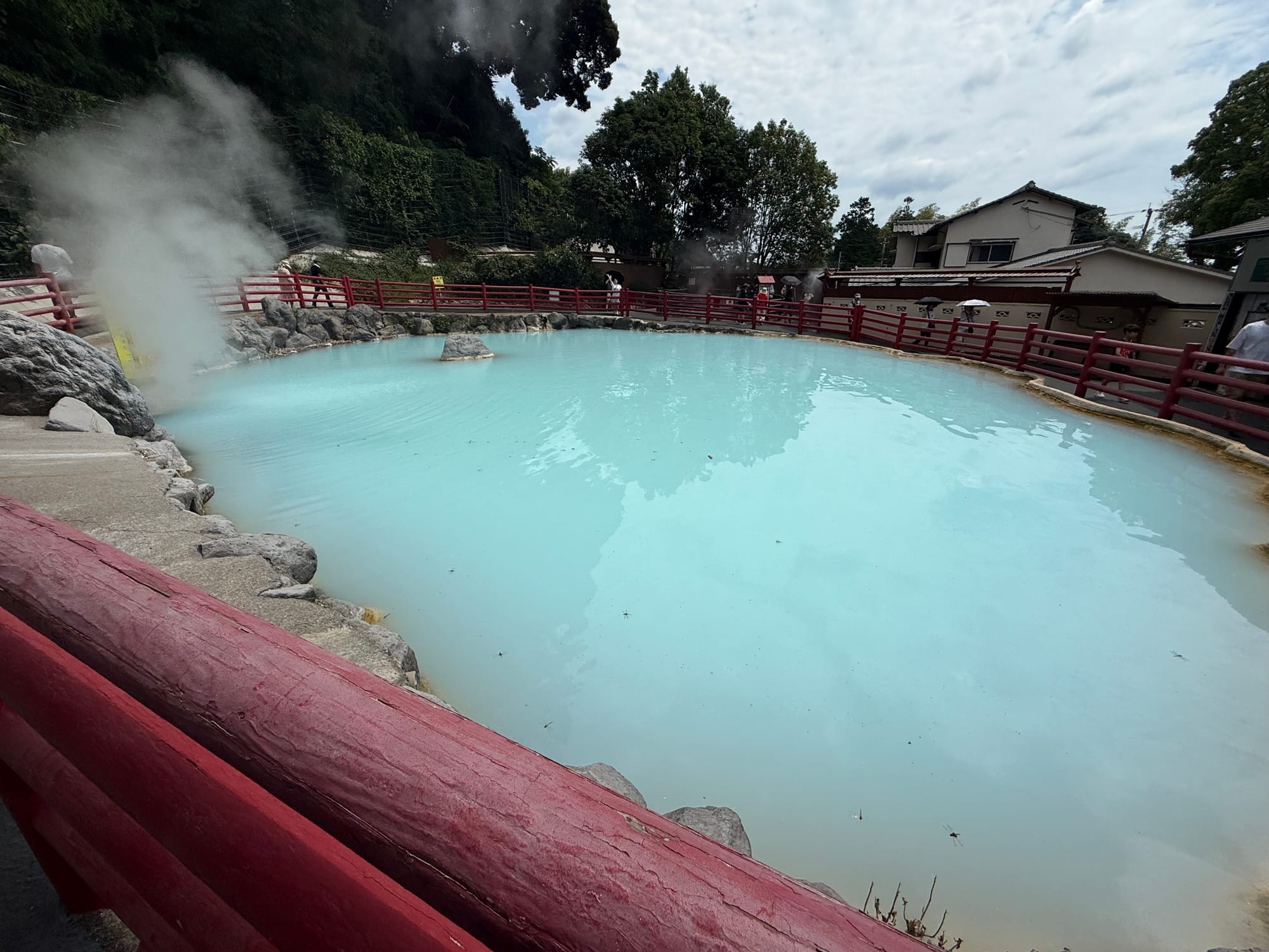 Milky blue geothermal pond at Kamado Jigoku in Beppu, surrounded by red railings, rocks and rising steam, with mineral-rich water creating an almost opaque appearance.