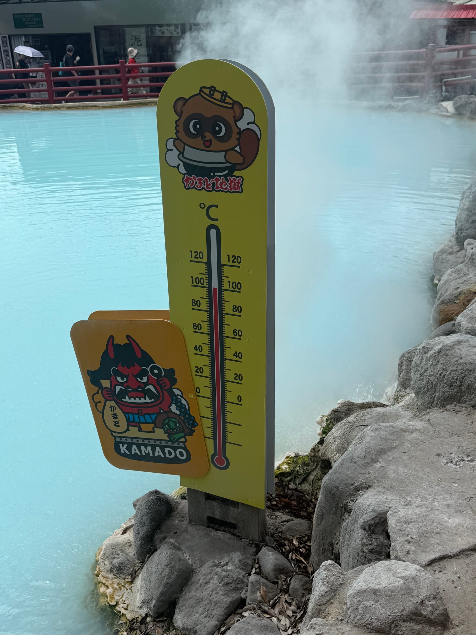 Thermometer beside a steaming blue geothermal pond at Kamado Jigoku in Beppu, showing the high temperature of the mineral-rich hot spring water.