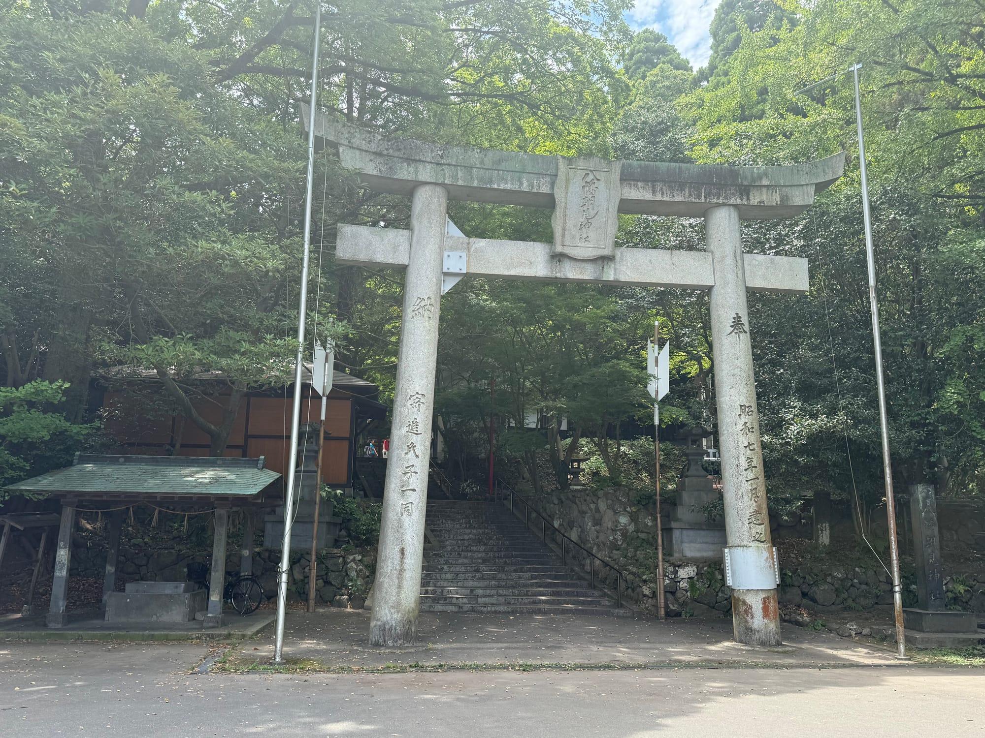 Stone torii gate at Hachiman Asami Shrine in Beppu, standing before a flight of steps leading into a wooded shrine precinct shaded by mature trees.