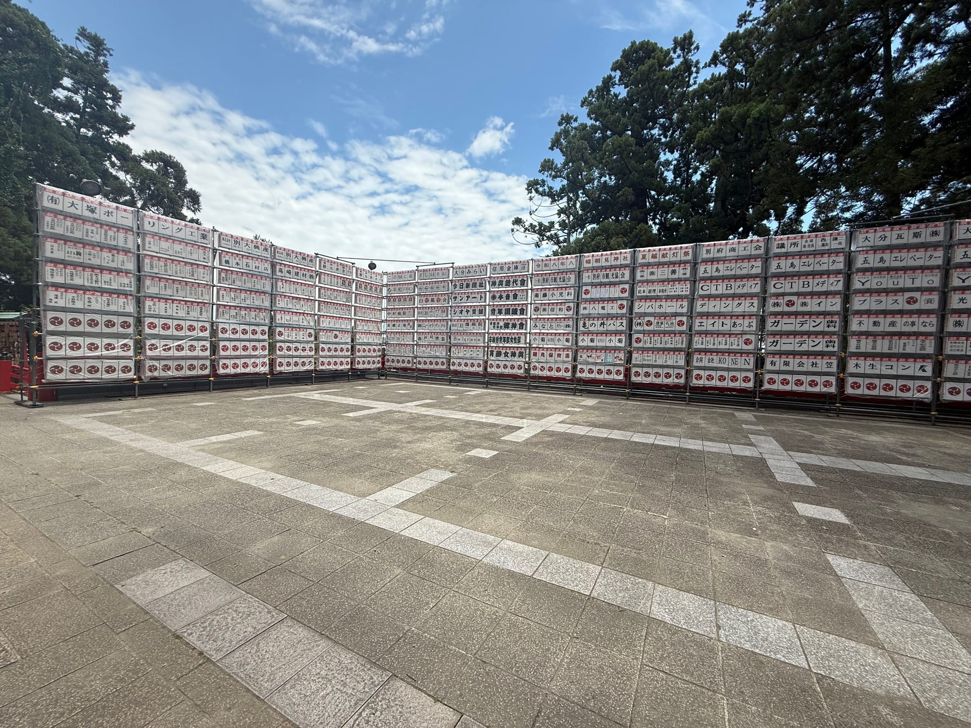 Rows of white lanterns and sponsor boards displayed in the grounds of Hachiman Asami Shrine in Beppu, set beneath tall trees and a bright summer sky.