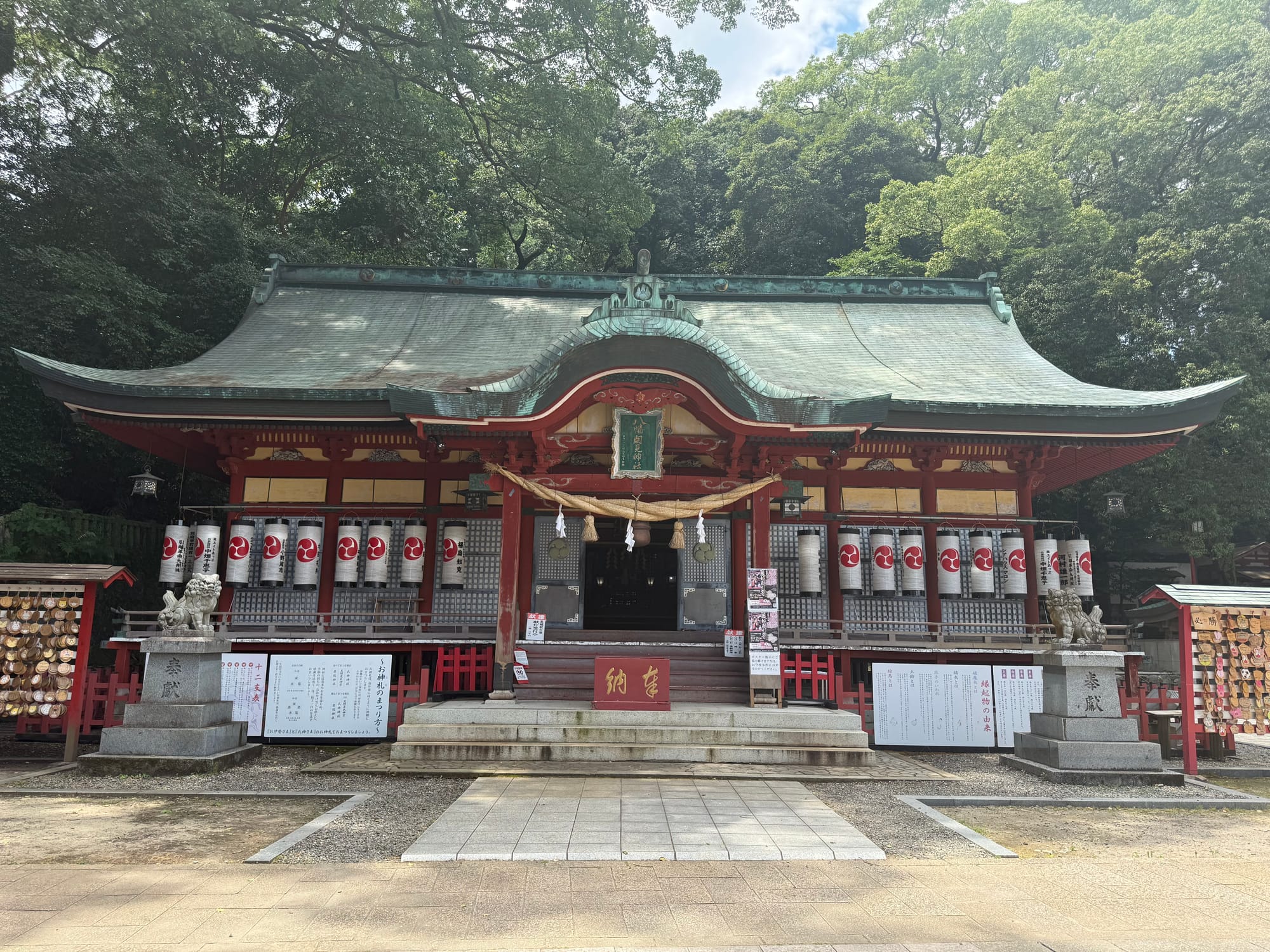 Main hall of Hachiman Asami Shrine in Beppu, featuring red-painted wooden architecture, lanterns, guardian statues and a copper-green roof beneath towering trees.