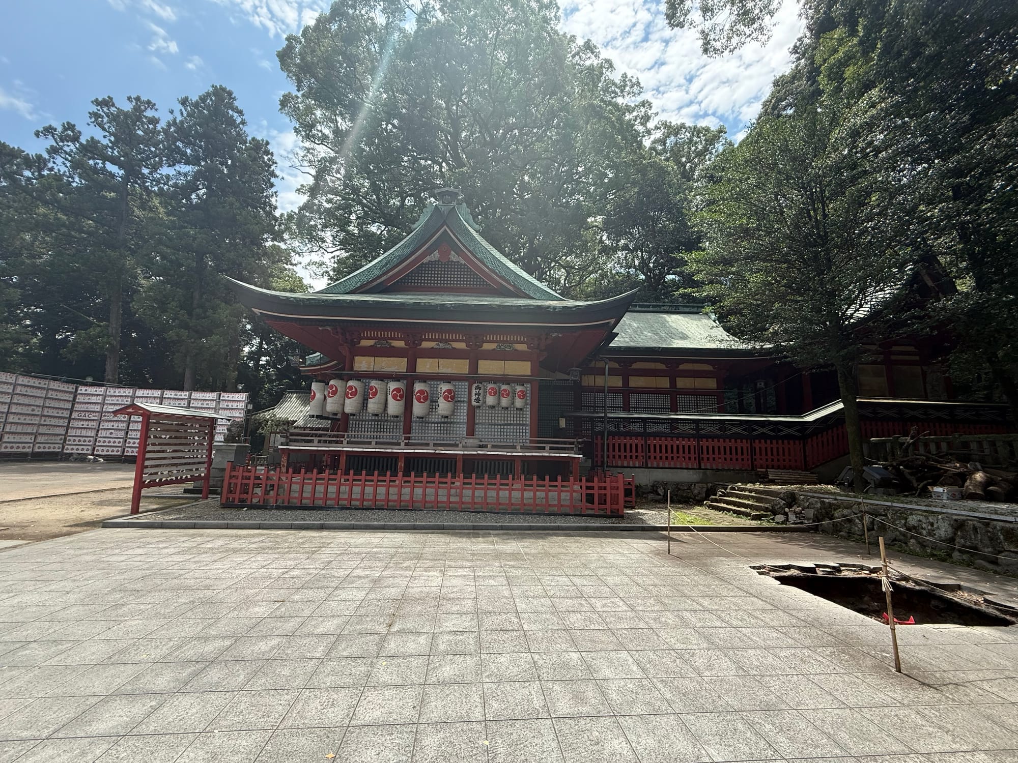 Side view of the main hall of Hachiman Asami Shrine in Beppu, featuring red-painted wooden architecture, lanterns, a copper-green roof and towering trees.