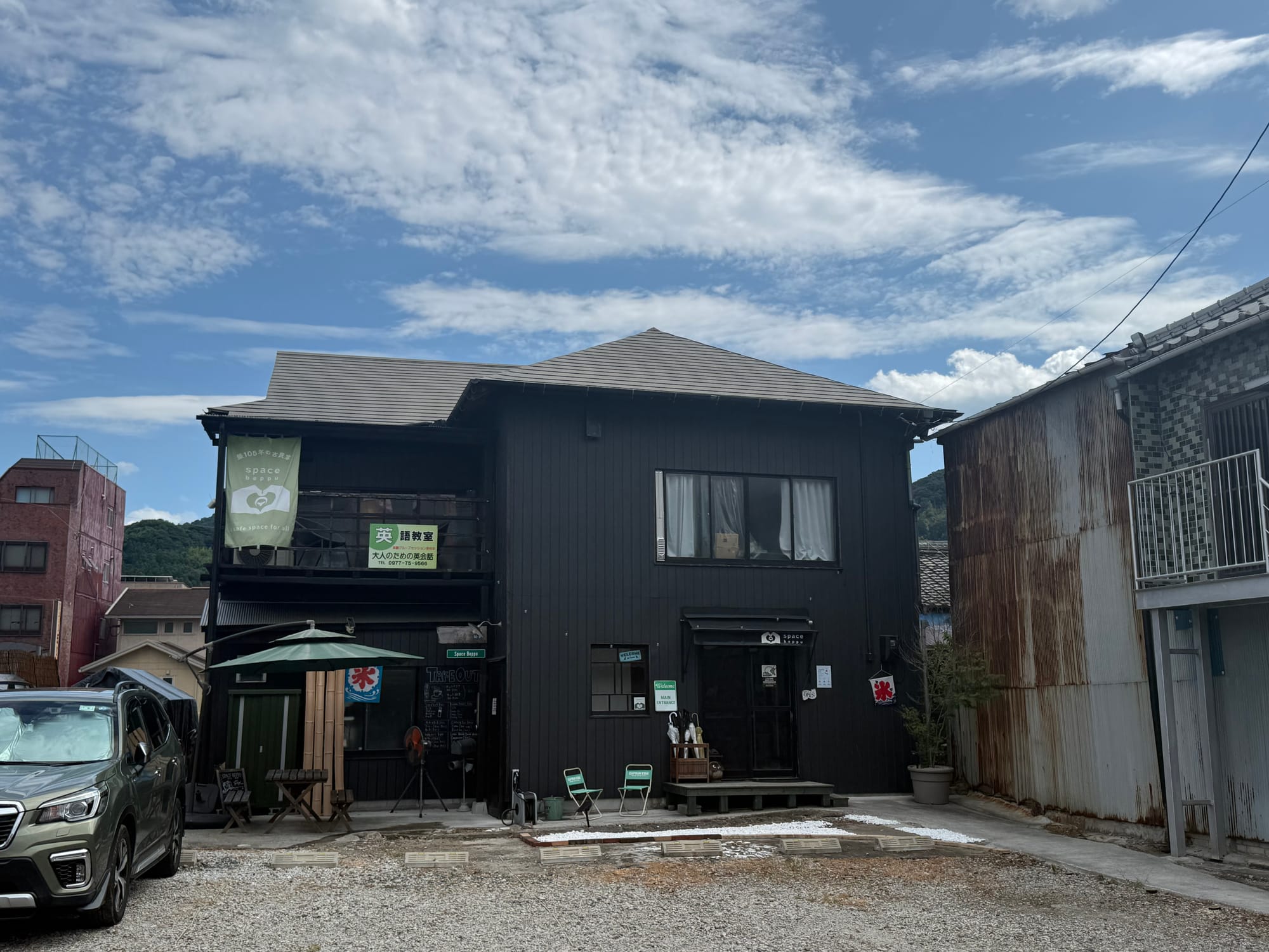 Traditional dark wooden building housing Space Beppu in central Beppu, with signage, seating outside and surrounding historic streets beneath a bright summer sky.