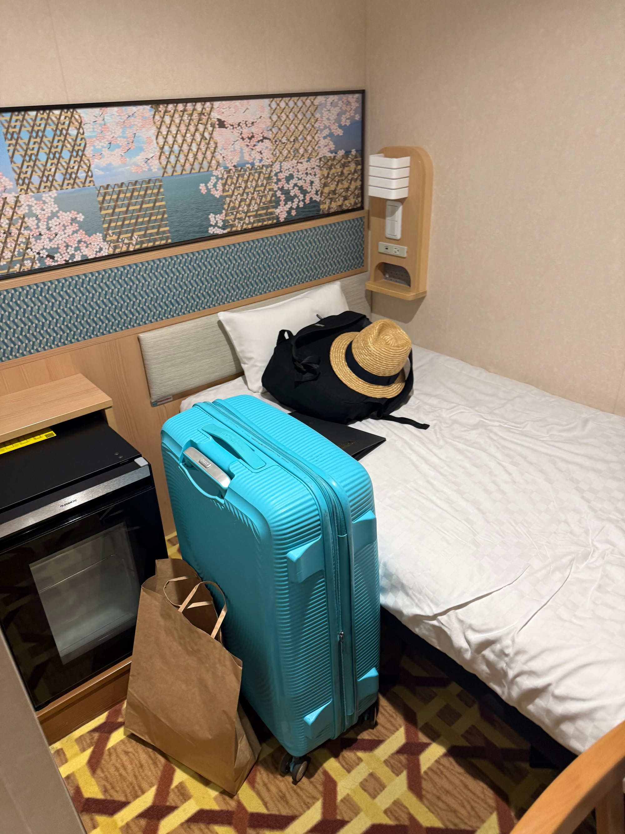 Compact private cabin aboard the Sunflower Ferry, featuring a single bed, luggage, refrigerator, storage unit and decorative wall panel inspired by cherry blossom designs.