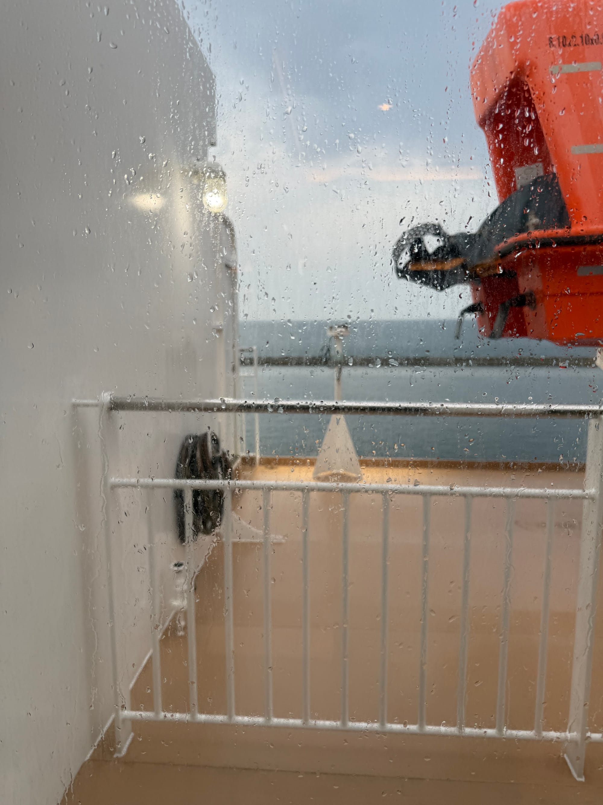 Rain-covered window on the Sunflower Ferry overlooking an outside deck, with an orange lifeboat, grey sea and overcast skies visible through droplets on the glass.