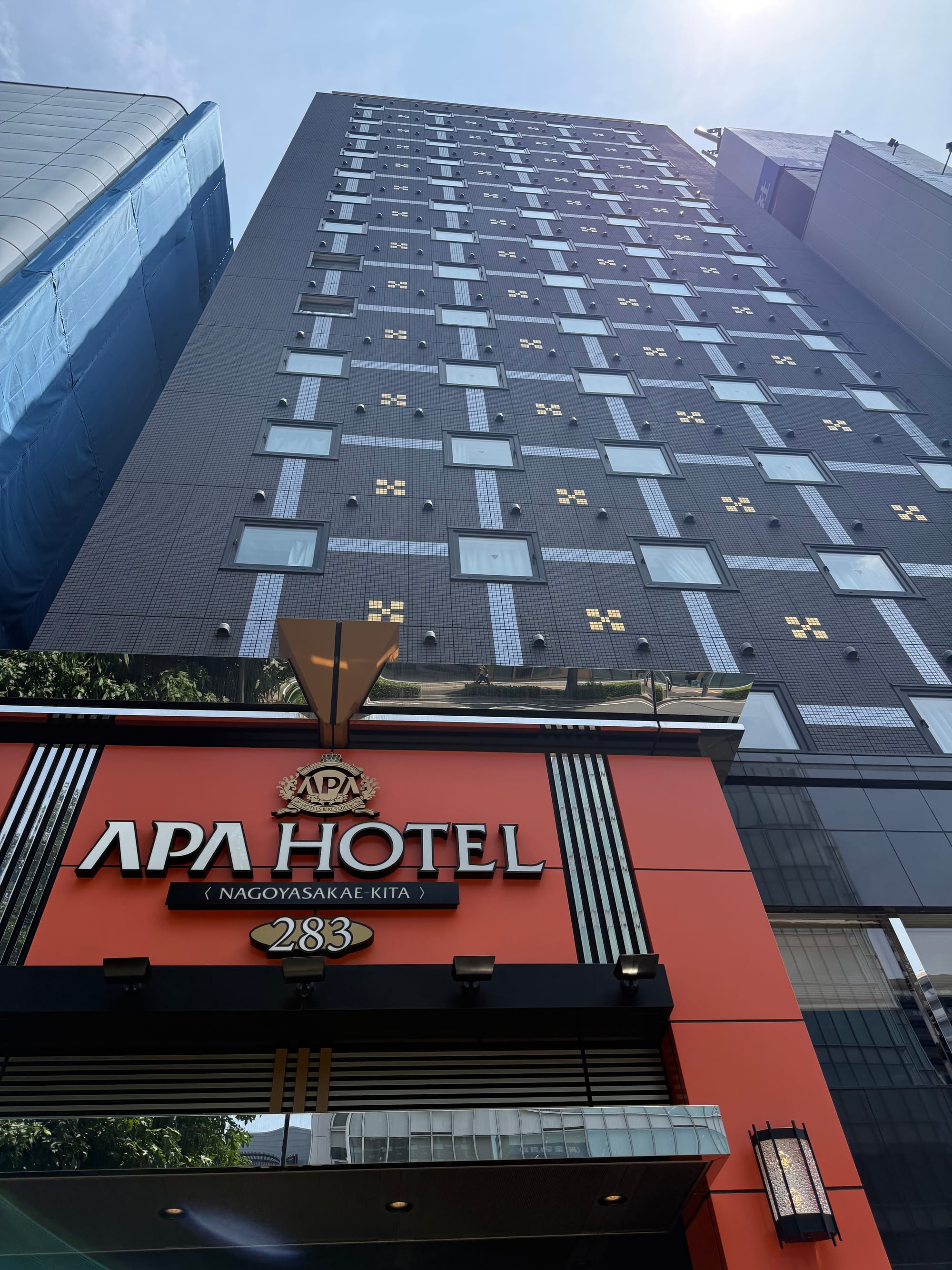 APA Hotel Nagoya Sakae-Kita, a modern high-rise hotel viewed from street level, with its distinctive orange entrance sign beneath a clear summer sky.