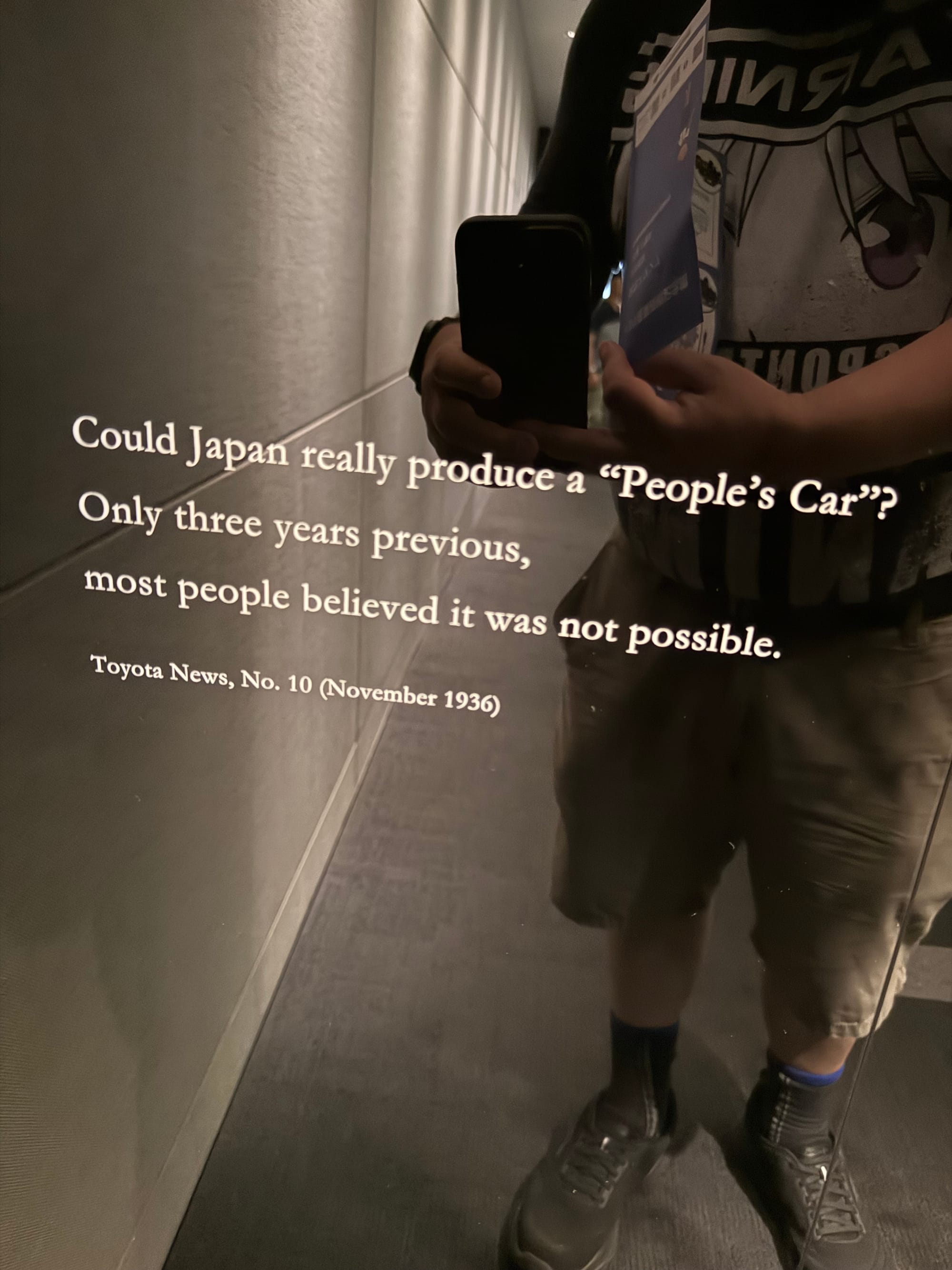 Exhibit at the Toyota Commemorative Museum featuring a 1936 quote questioning whether Japan could produce a "people's car", reflected in a mirrored display panel.
