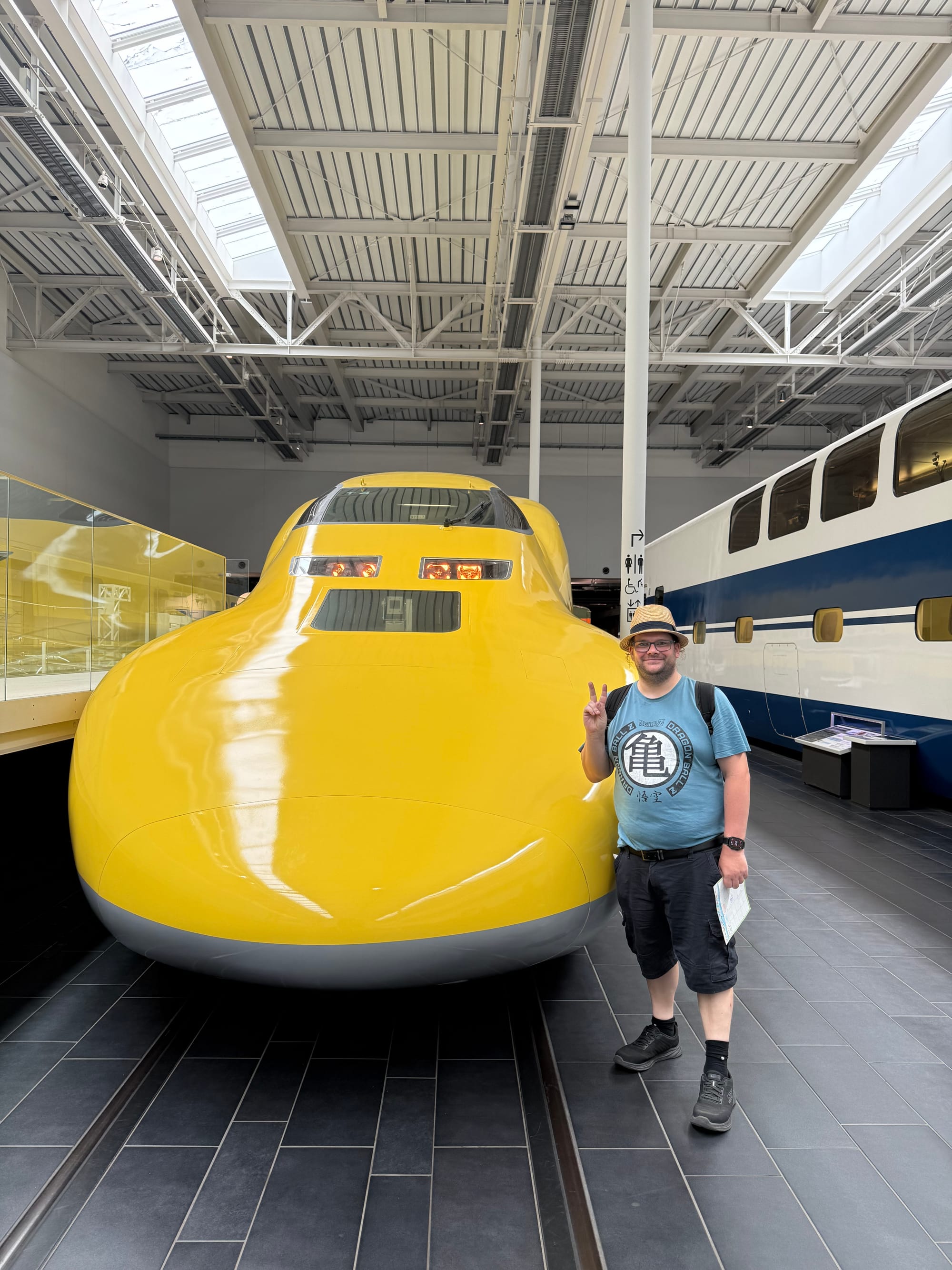 Visitor poses beside Doctor Yellow at the SCMAGLEV and Railway Park in Nagoya, making a peace sign in front of the iconic yellow Shinkansen inspection train.