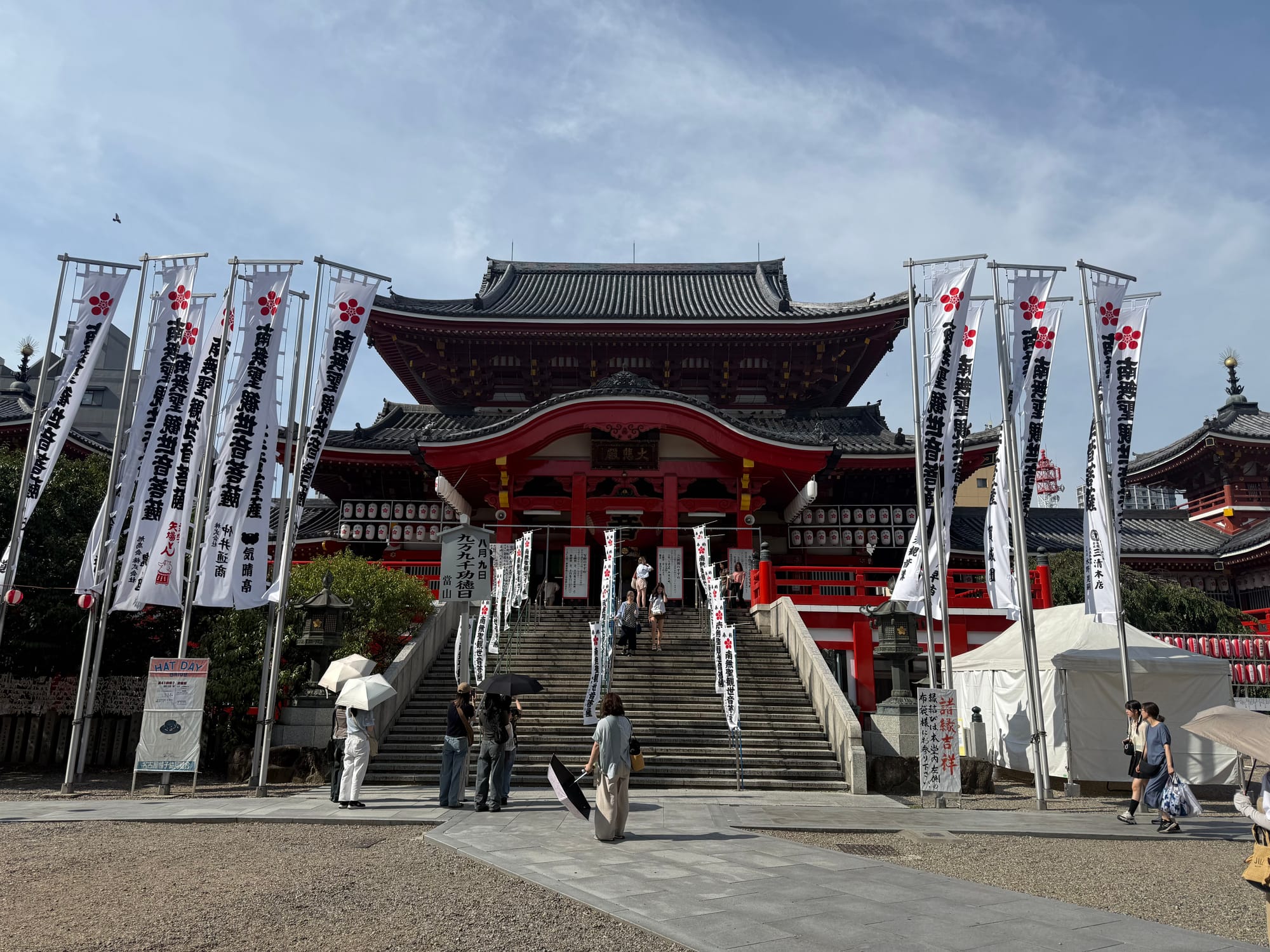 Main hall of Ōsu Kannon Temple in Nagoya, featuring striking red-and-white architecture, stone steps, festival banners and visitors gathered beneath a bright summer sky.