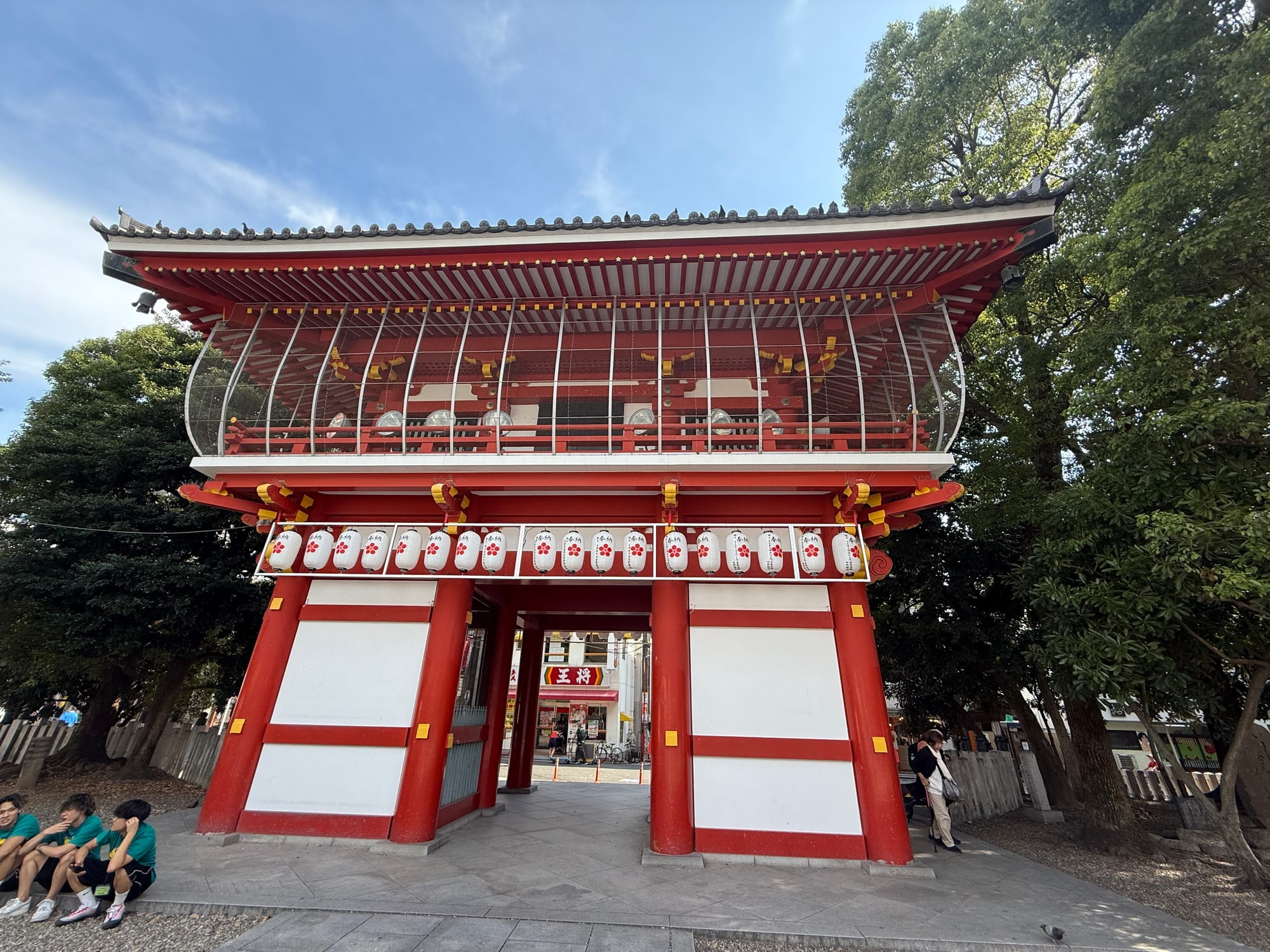 Red-and-white temple gate at Ōsu Kannon Temple in Nagoya, decorated with rows of lanterns and traditional architectural details, framed by mature trees.