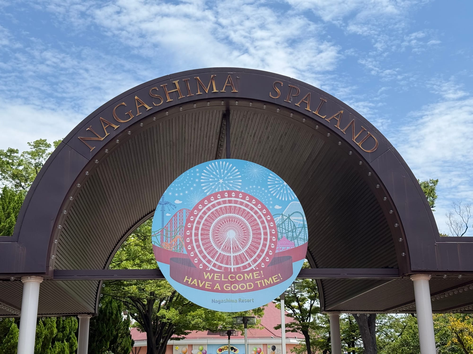 Entrance arch at Nagashima Spa Land, featuring a welcome sign with illustrations of roller coasters and a Ferris wheel beneath a bright blue sky with scattered clouds.