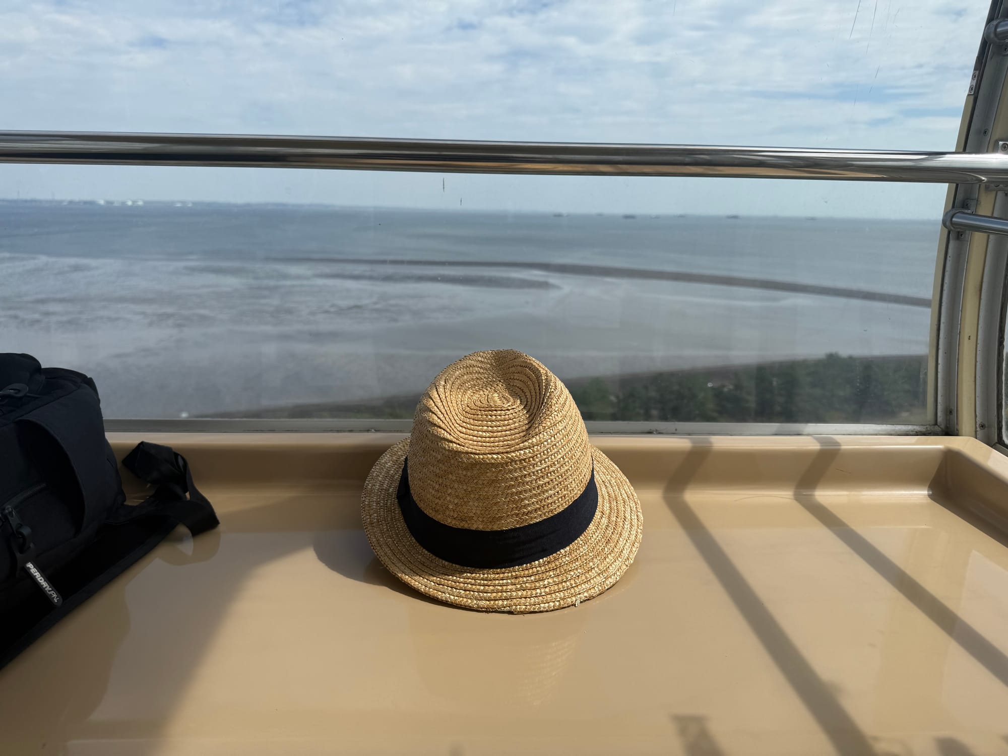 Straw hat with a black band resting on a Ferris wheel cabin ledge at Nagashima Spa Land, overlooking tidal flats, waterways and the distant shoreline beneath clear skies.