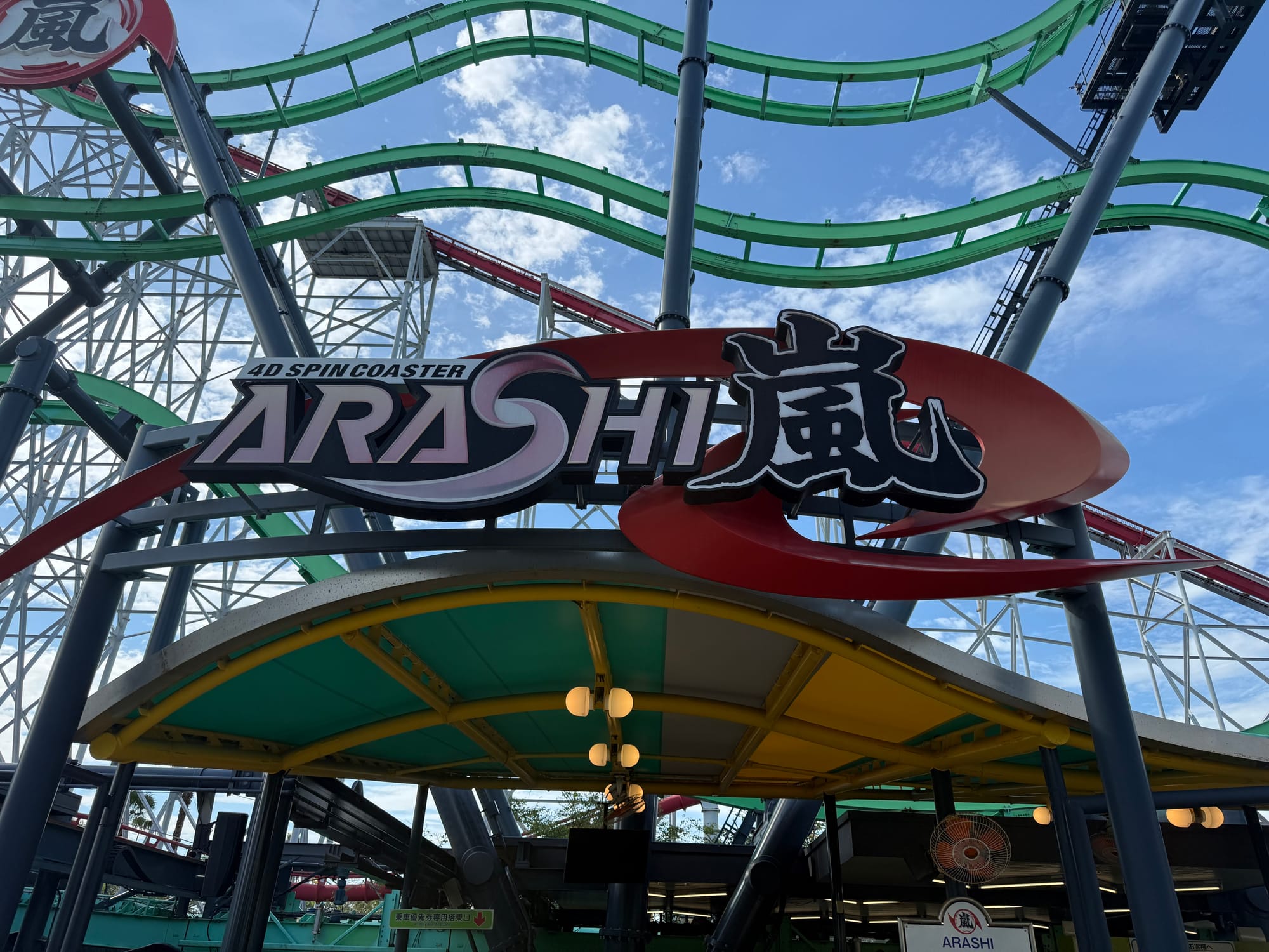 Entrance sign for Arashi, a 4D Spin Coaster at Nagashima Spa Land, with green track weaving overhead and the towering structure of Steel Dragon 2000 visible behind.