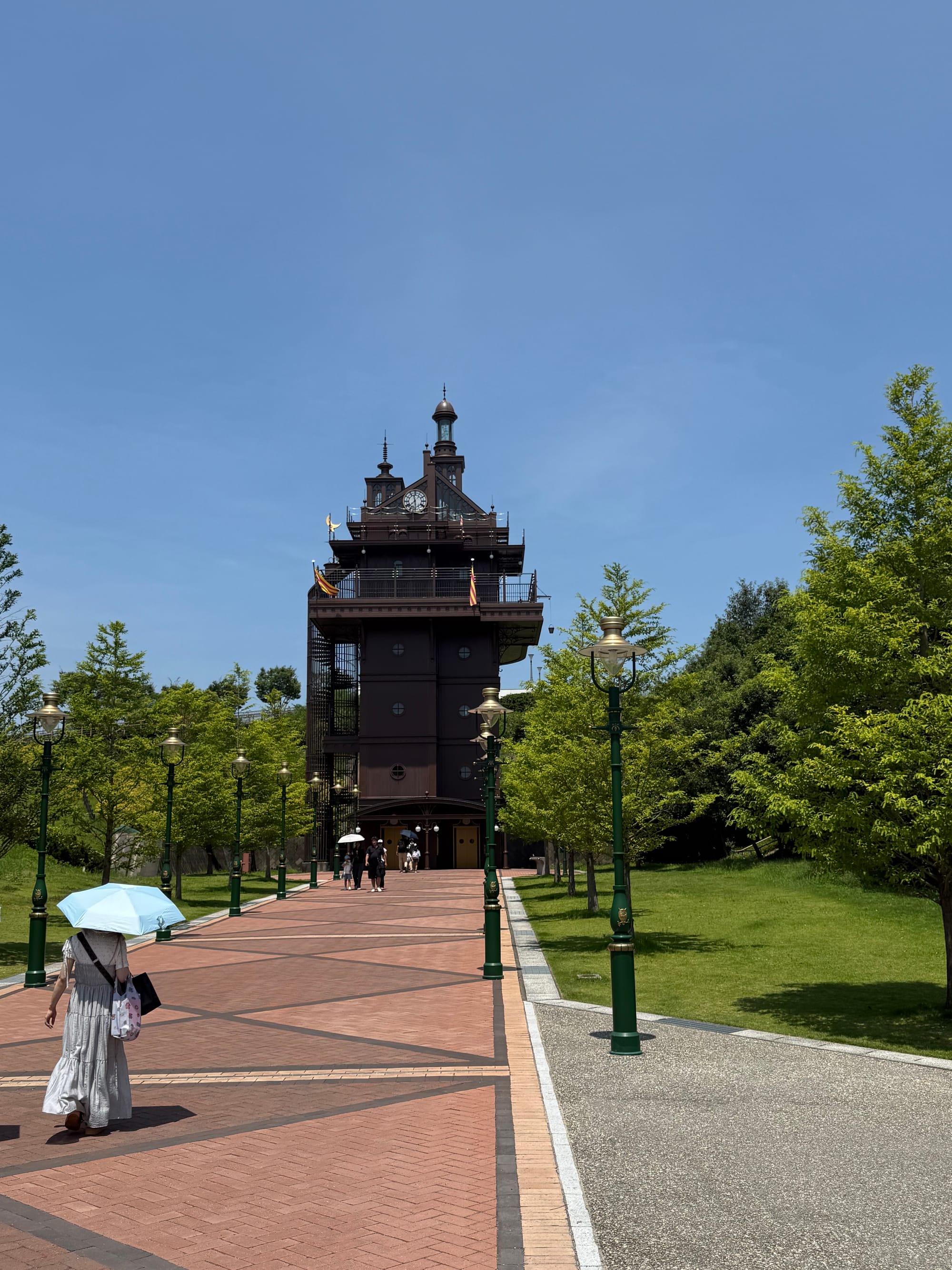 The Elevator Tower at Ghibli Park, inspired by the architecture of Studio Ghibli films, stands at the end of a tree-lined promenade beneath a clear summer sky.