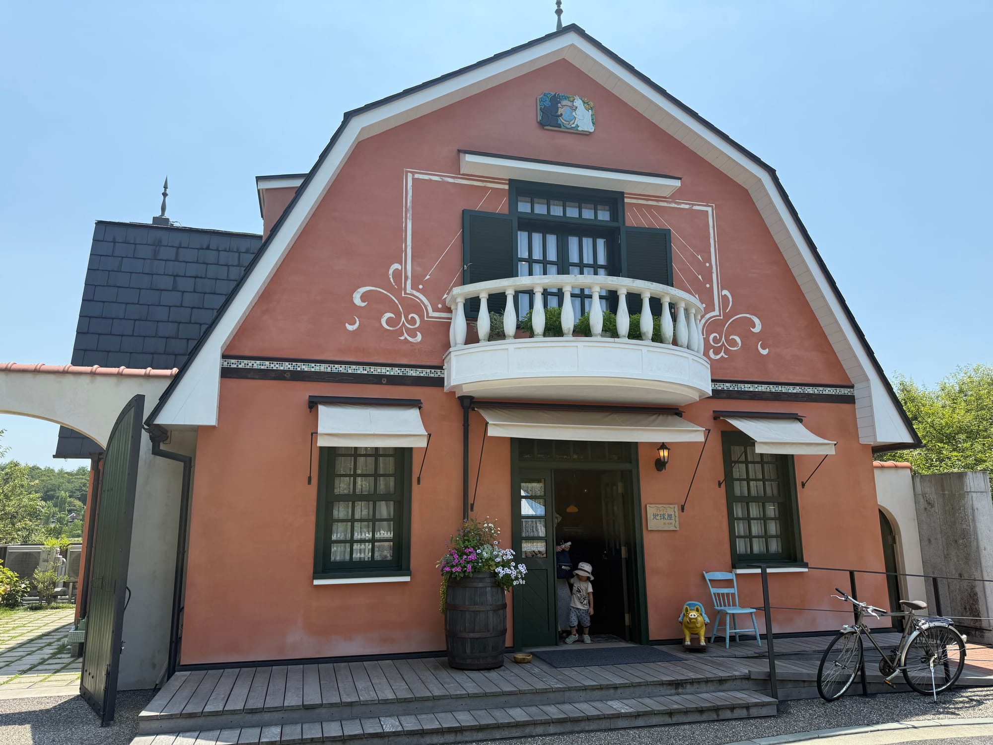 World Emporium building at Ghibli Park, featuring peach-coloured walls, a balcony, green shutters, flower displays and a bicycle outside the entrance on a sunny day.