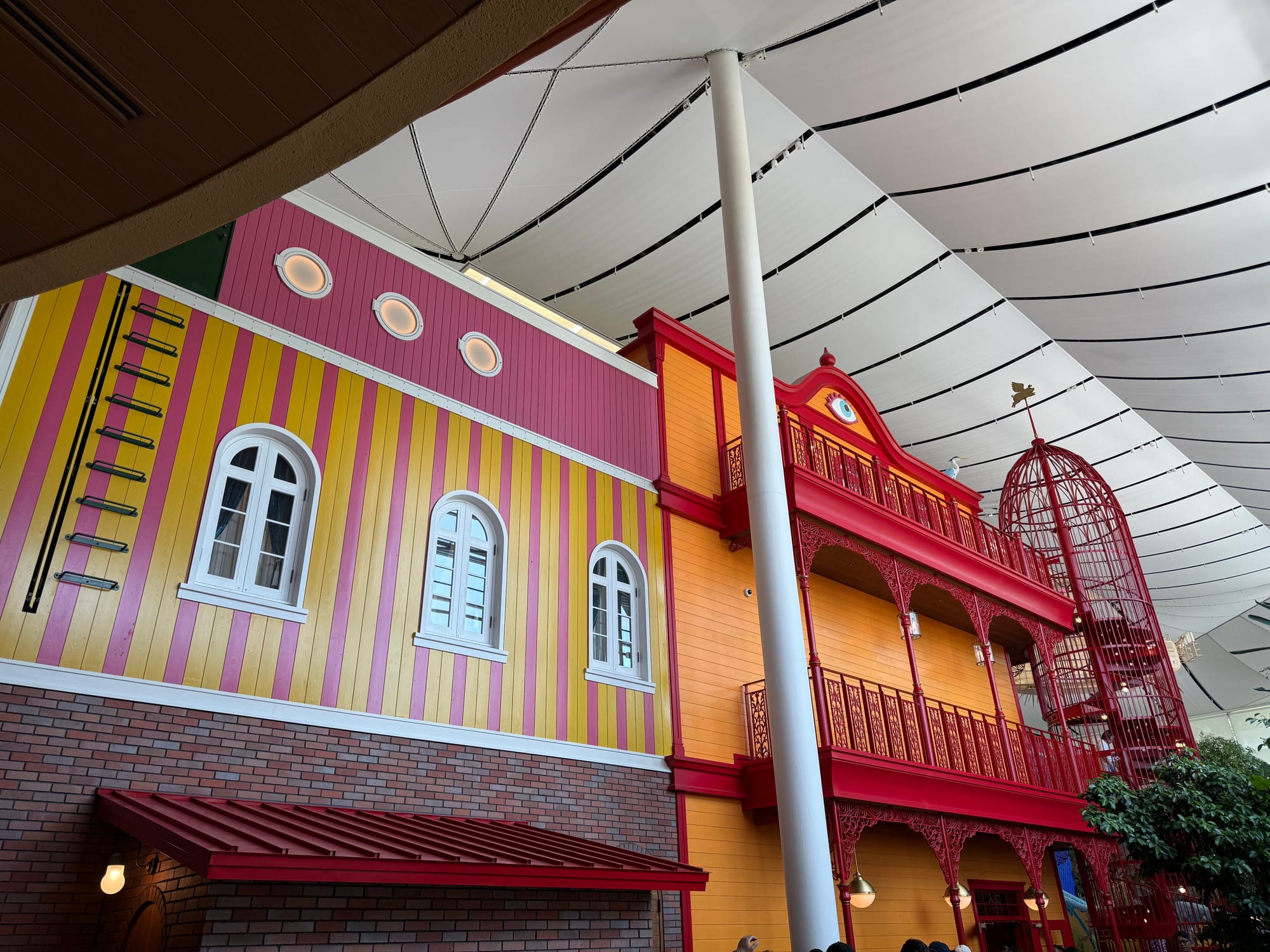 Colourful interior of the Grand Warehouse at Ghibli Park, featuring whimsical building façades, ornate red balconies, a giant birdcage and a sweeping white canopy roof.