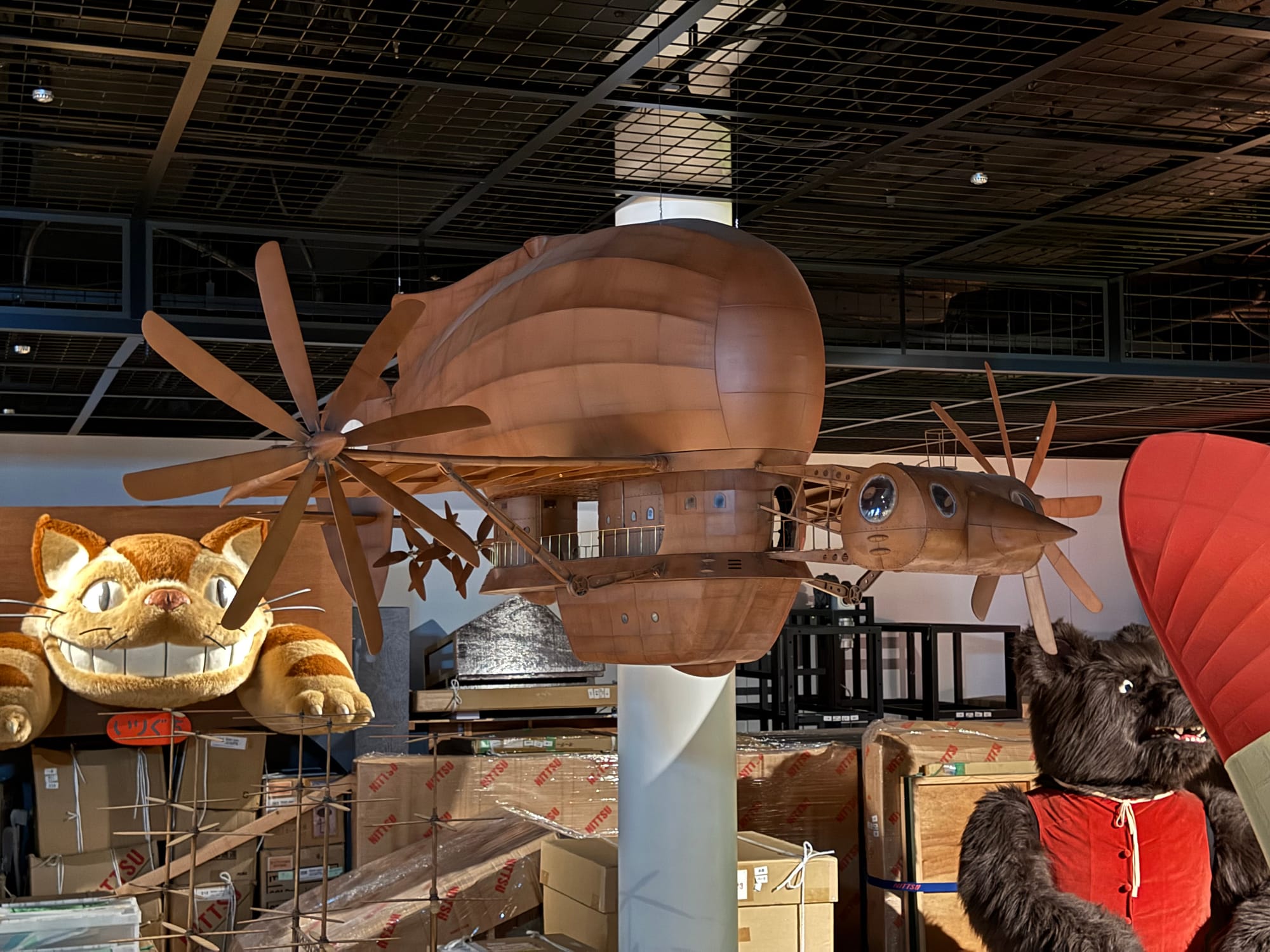 Model of the flying machine from Castle in the Sky displayed inside Ghibli Park's Grand Warehouse, surrounded by props including the Catbus and other Studio Ghibli exhibits.