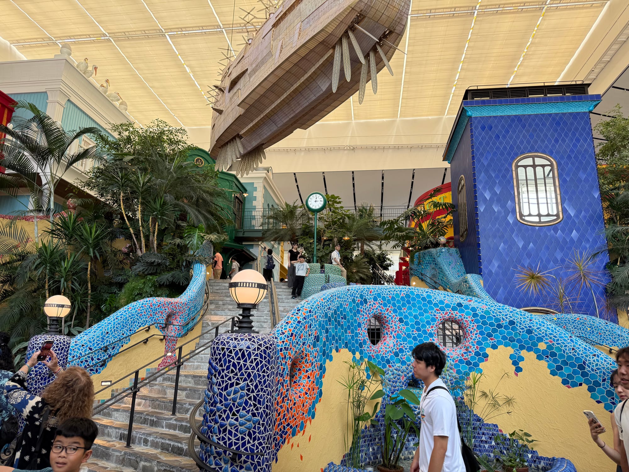 The Grand Staircase inside Ghibli Park's Grand Warehouse, featuring colourful mosaic-covered railings, lush tropical plants and a large airship suspended overhead.