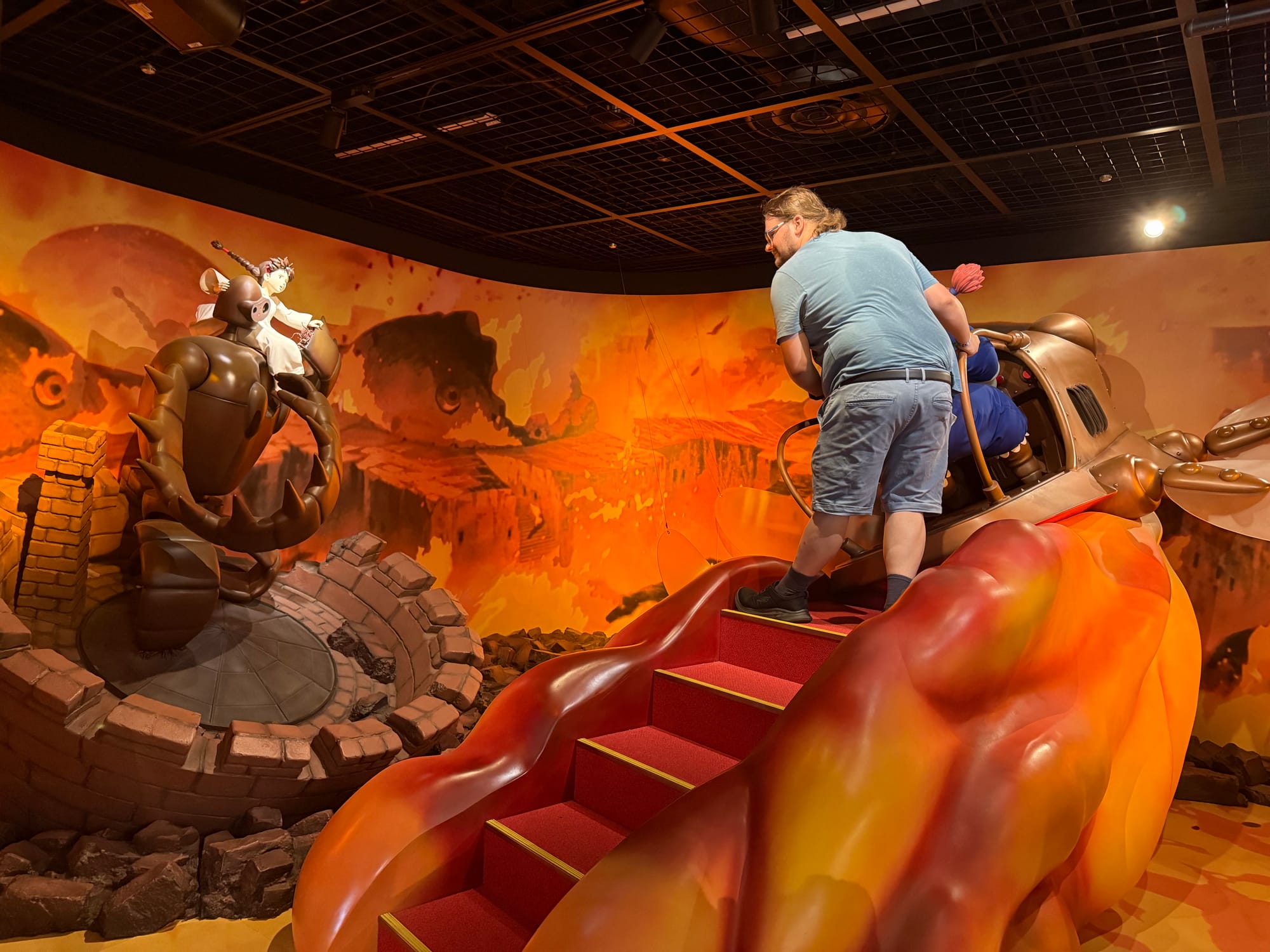 Visitor boards Dola's flaptor from Castle in the Sky in the Becoming Characters in Memorable Ghibli Scenes exhibit, with Sheeta riding a Laputian robot soldier nearby.