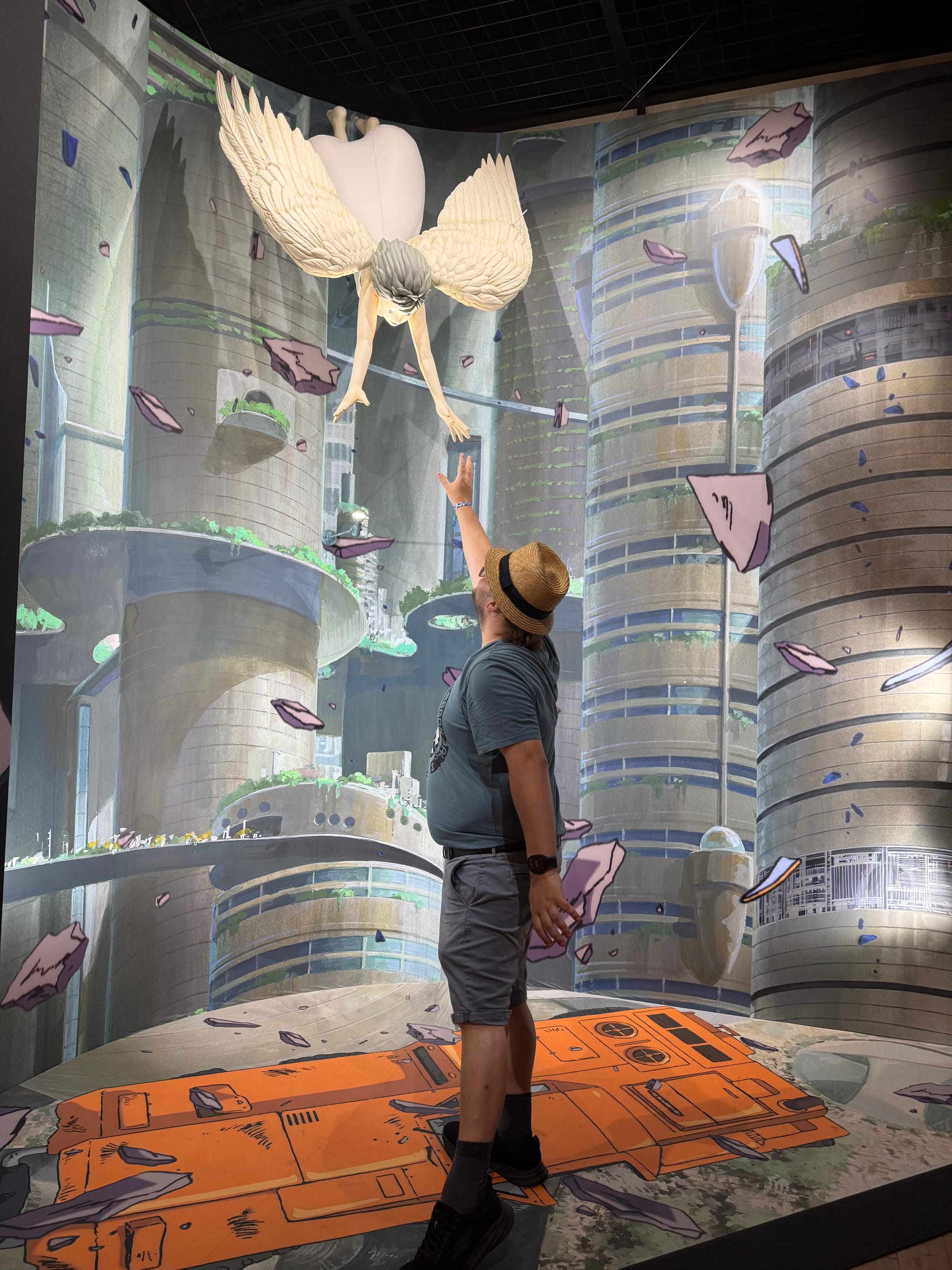 Visitor reaches towards the winged girl from On Your Mark in the Becoming Characters in Memorable Ghibli Scenes exhibit, set against a futuristic cityscape filled with floating debris.