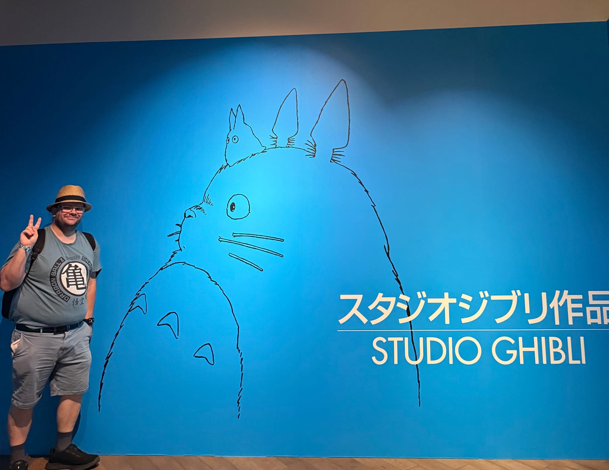 Visitor poses beside a large Studio Ghibli Totoro wall graphic in the Grand Warehouse at Ghibli Park, featuring a blue backdrop and line-art illustration of Totoro.