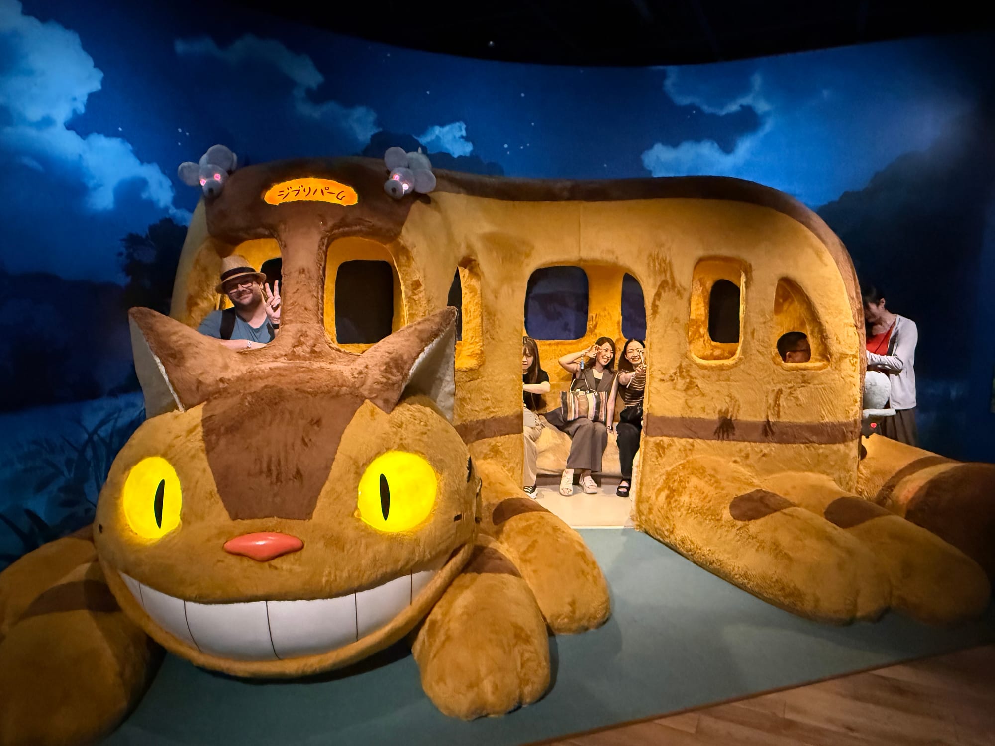 Visitor poses inside a full-size Catbus at Ghibli Park's Grand Warehouse, with glowing yellow eyes, oversized fur-covered seats and a night-time forest backdrop.