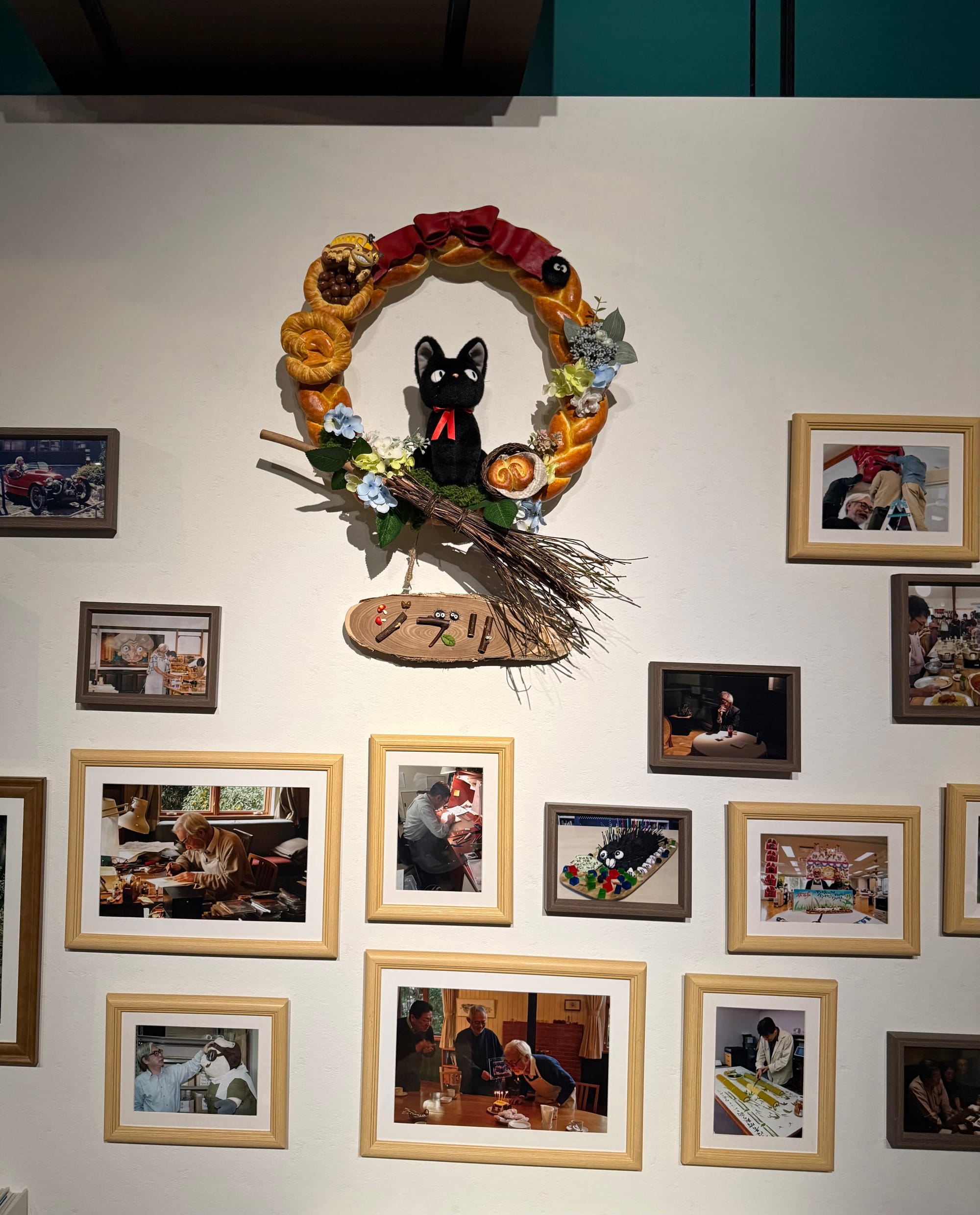 Jiji display from Kiki's Delivery Service inside Ghibli Park's Grand Warehouse, featuring the black cat perched on a broom within a decorative wreath surrounded by production photographs.