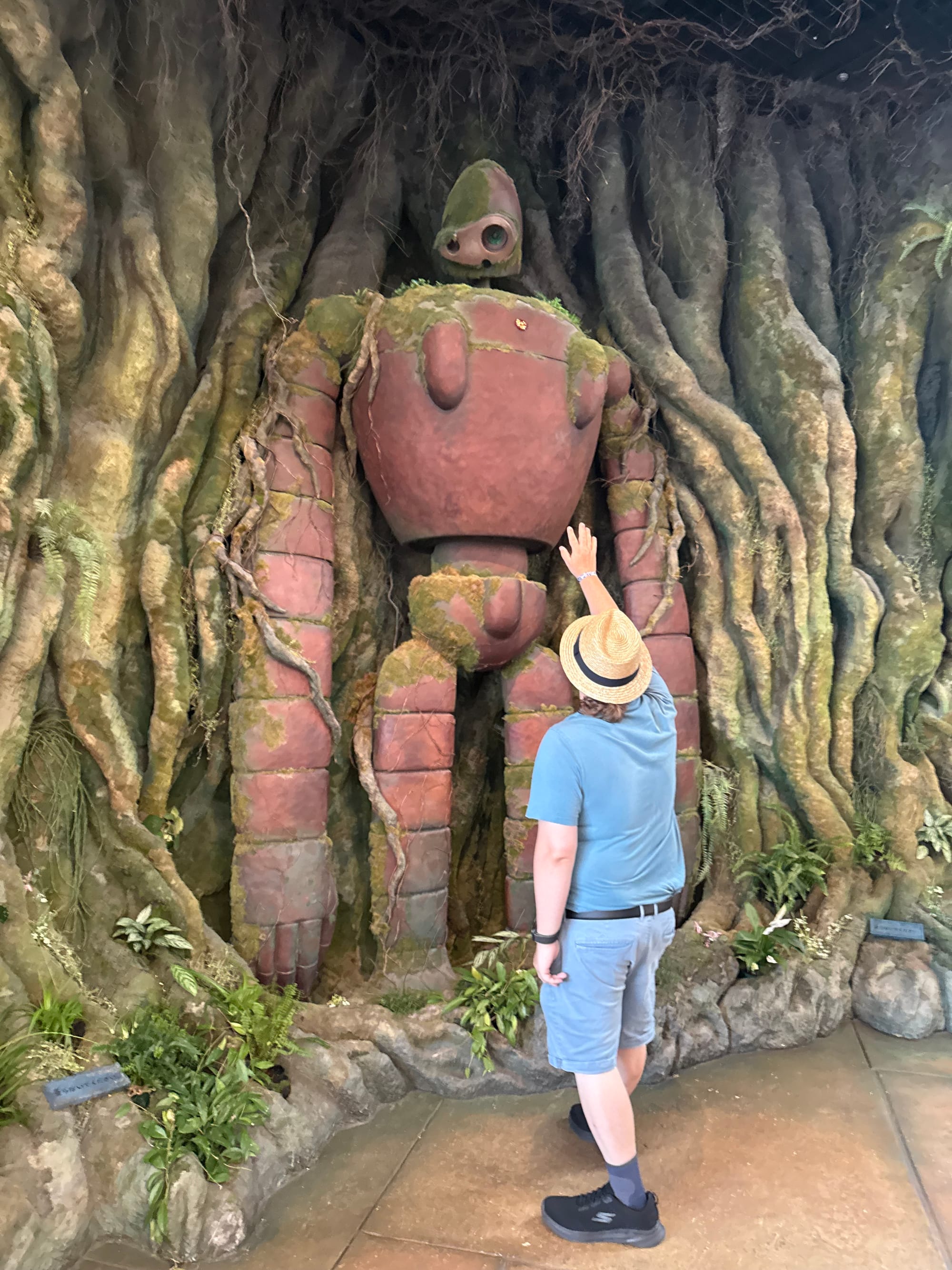 Visitor reaches towards a Laputian robot soldier from Castle in the Sky at Ghibli Park's Grand Warehouse, standing beneath giant tree roots and moss-covered stonework.