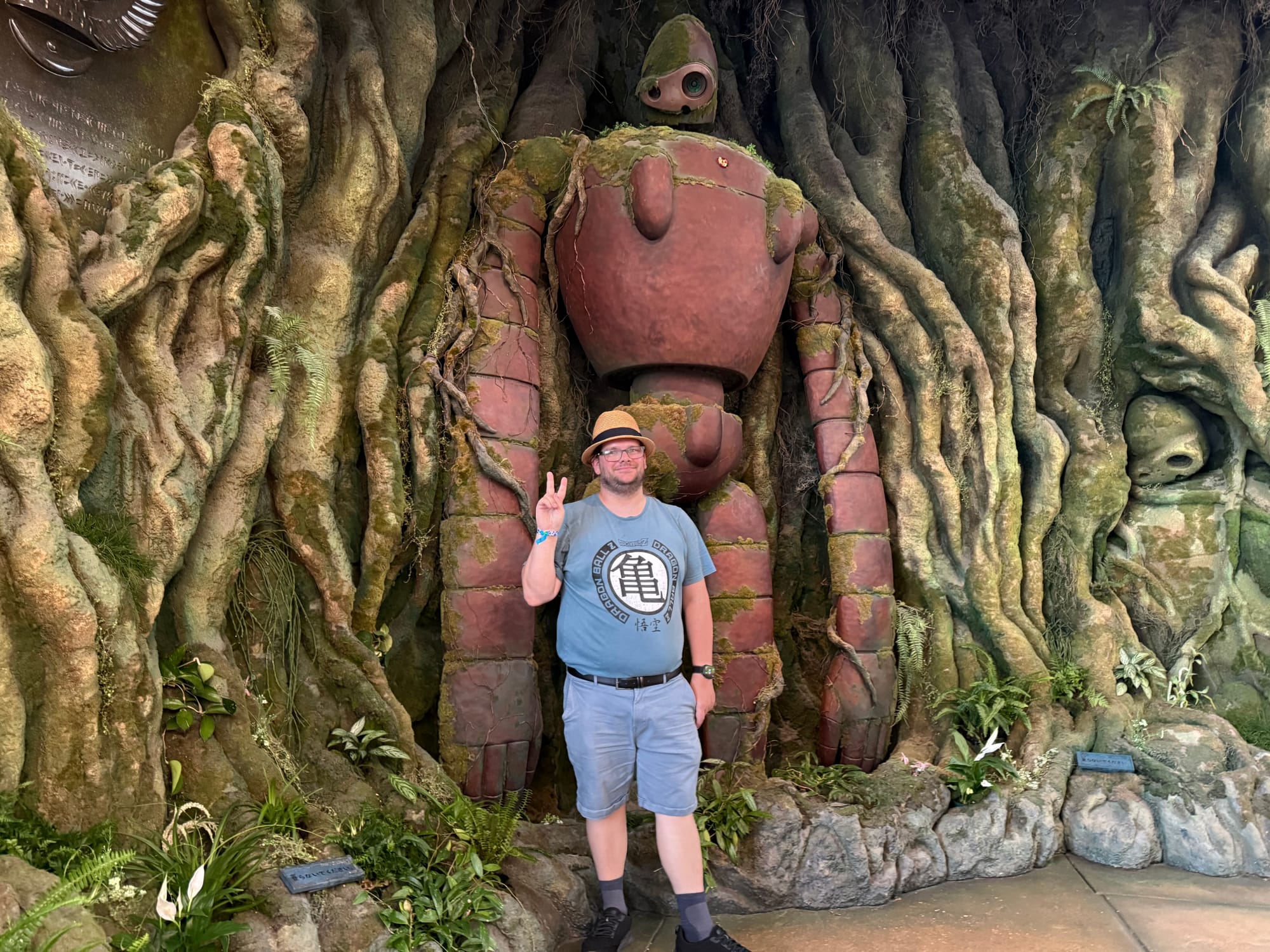 Visitor poses in front of a Laputian robot soldier from Castle in the Sky at Ghibli Park's Grand Warehouse, surrounded by giant tree roots, moss and forest-inspired scenery.