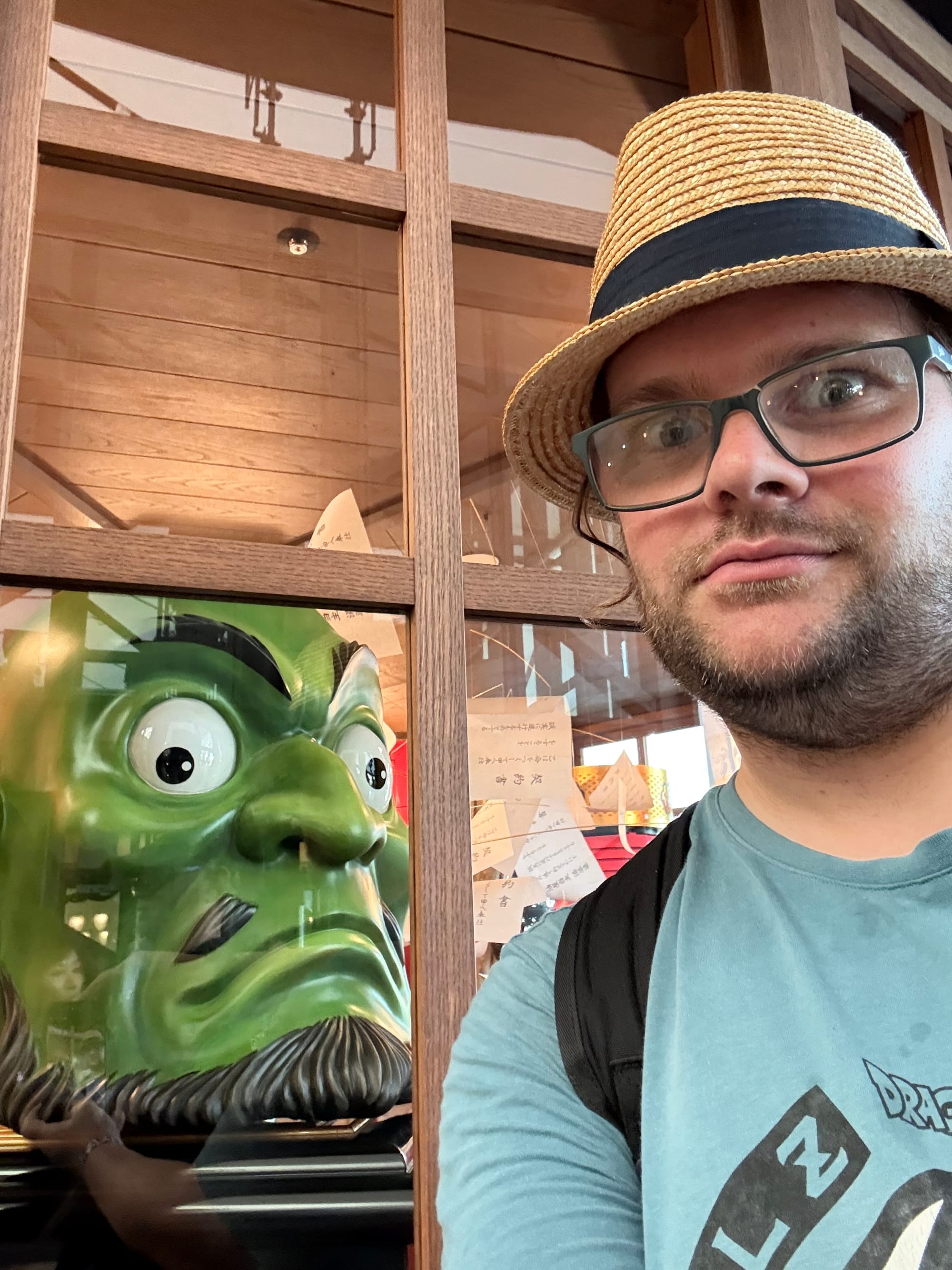 Visitor poses beside a large green Tengu mask display at Ghibli Park, with wooden-framed windows and numerous handwritten messages visible behind the glass.