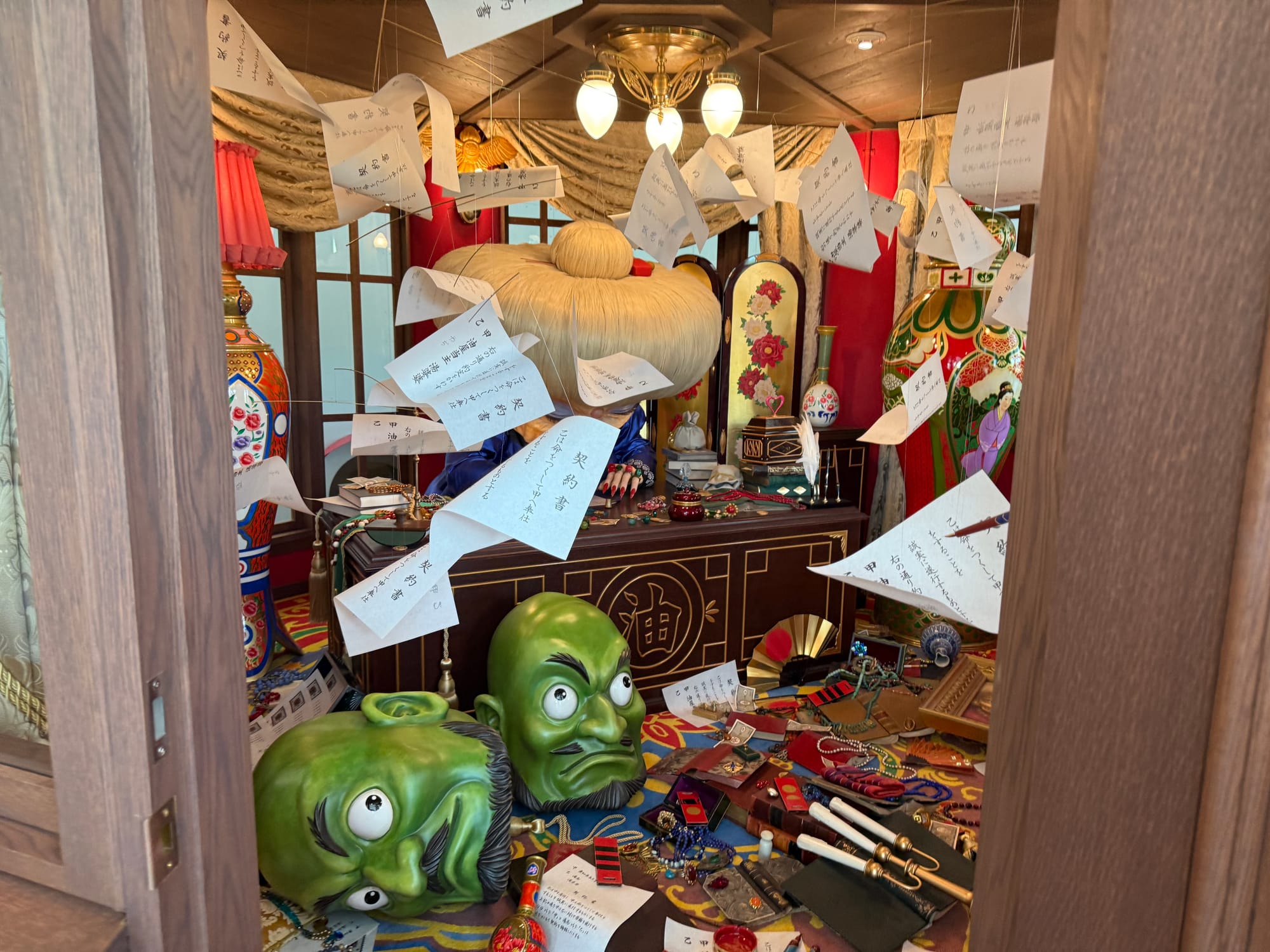 Display from Pom Poko inside Ghibli Park's Grand Warehouse, featuring giant tengu masks, scattered props, floating notes and a richly decorated room filled with folklore-inspired details.