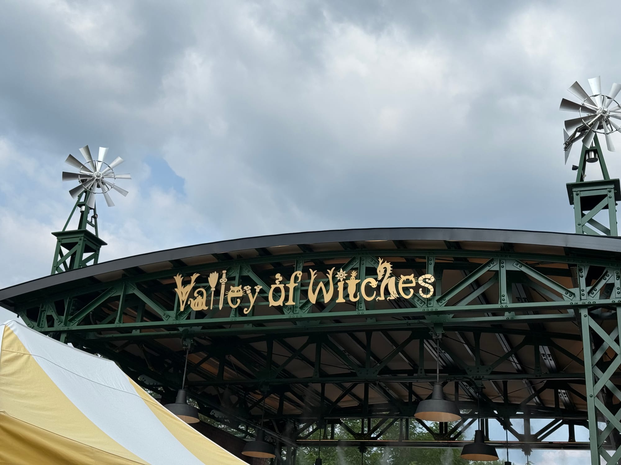 Entrance sign for the Valley of Witches at Ghibli Park, featuring ornate gold lettering on a green iron structure topped with decorative windmills beneath cloudy skies.