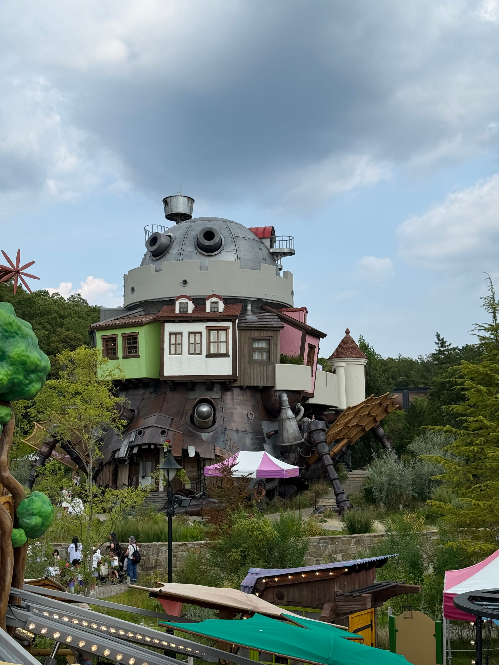 Howl's Moving Castle at Ghibli Park's Valley of Witches, featuring the towering mechanical castle with colourful rooms, metal legs, pipes and turrets beneath a cloudy sky.