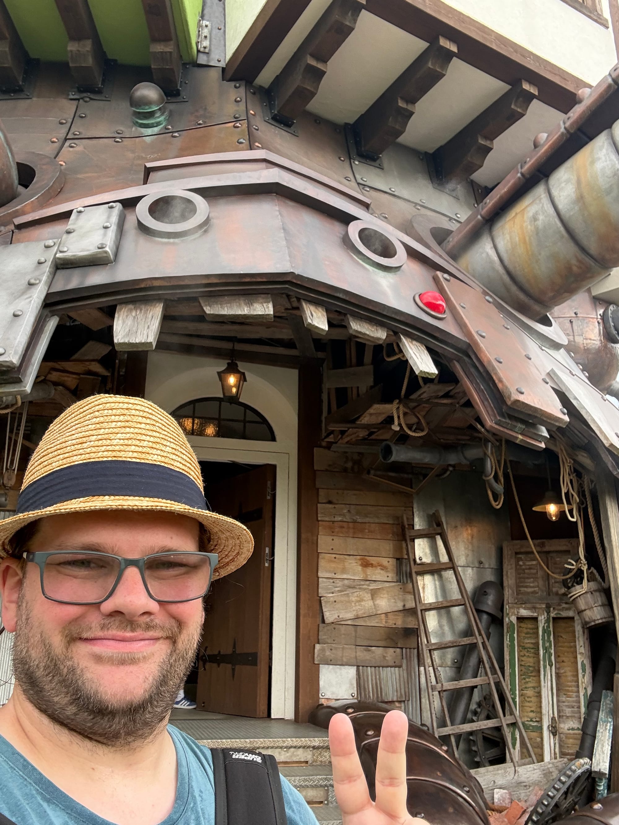 Visitor wearing a straw hat poses outside Howl's Moving Castle at Ghibli Park's Valley of Witches, with the castle's intricate pipes, metalwork and entrance behind.