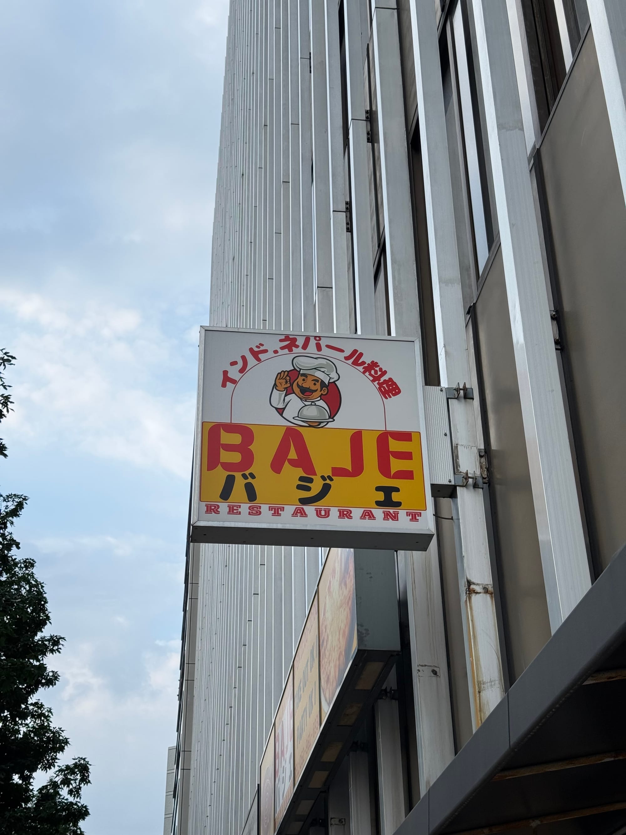 Sign for Baje Restaurant in Nagoya, featuring a smiling chef logo, Japanese text and bold yellow-and-red signage mounted on the side of a city-centre building.
