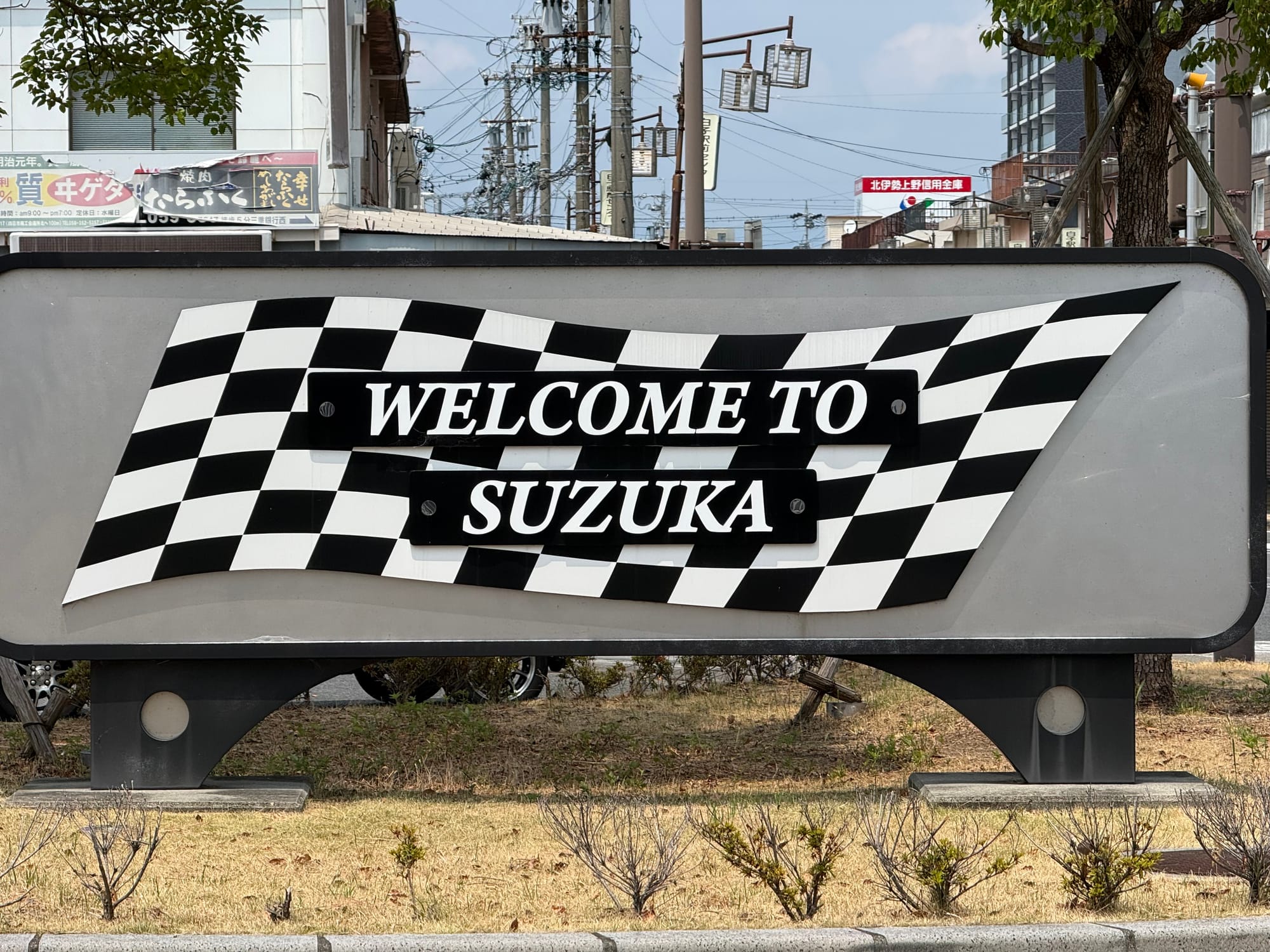 "Welcome to Suzuka" sign featuring a chequered flag design, displayed beside a road in Suzuka City, reflecting the area's strong association with motorsport.