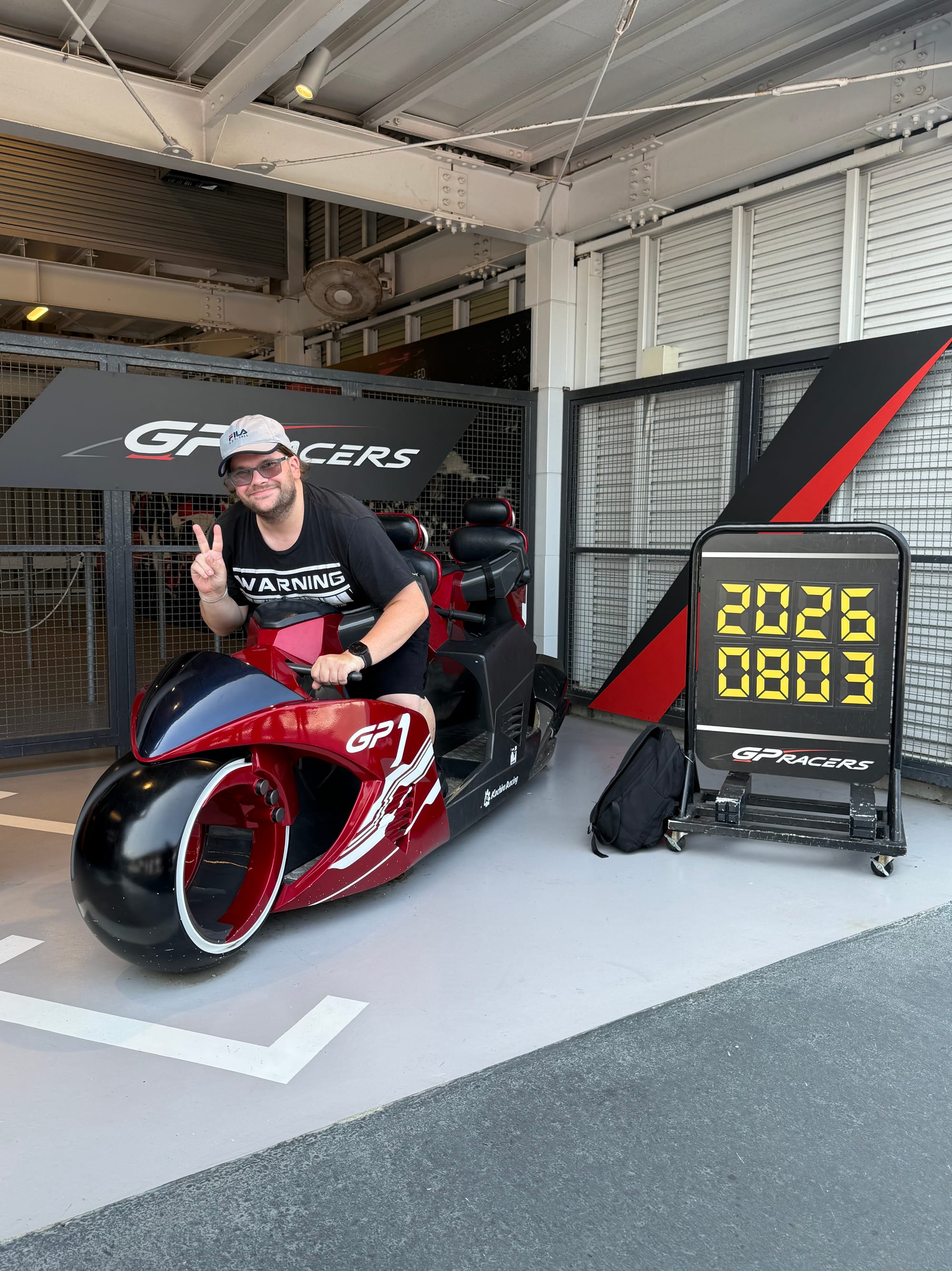 Visitor seated on a GP Racers motorcycle simulator at Suzuka Circuit Park, making a peace sign beside a board displaying the date, Japanese style..