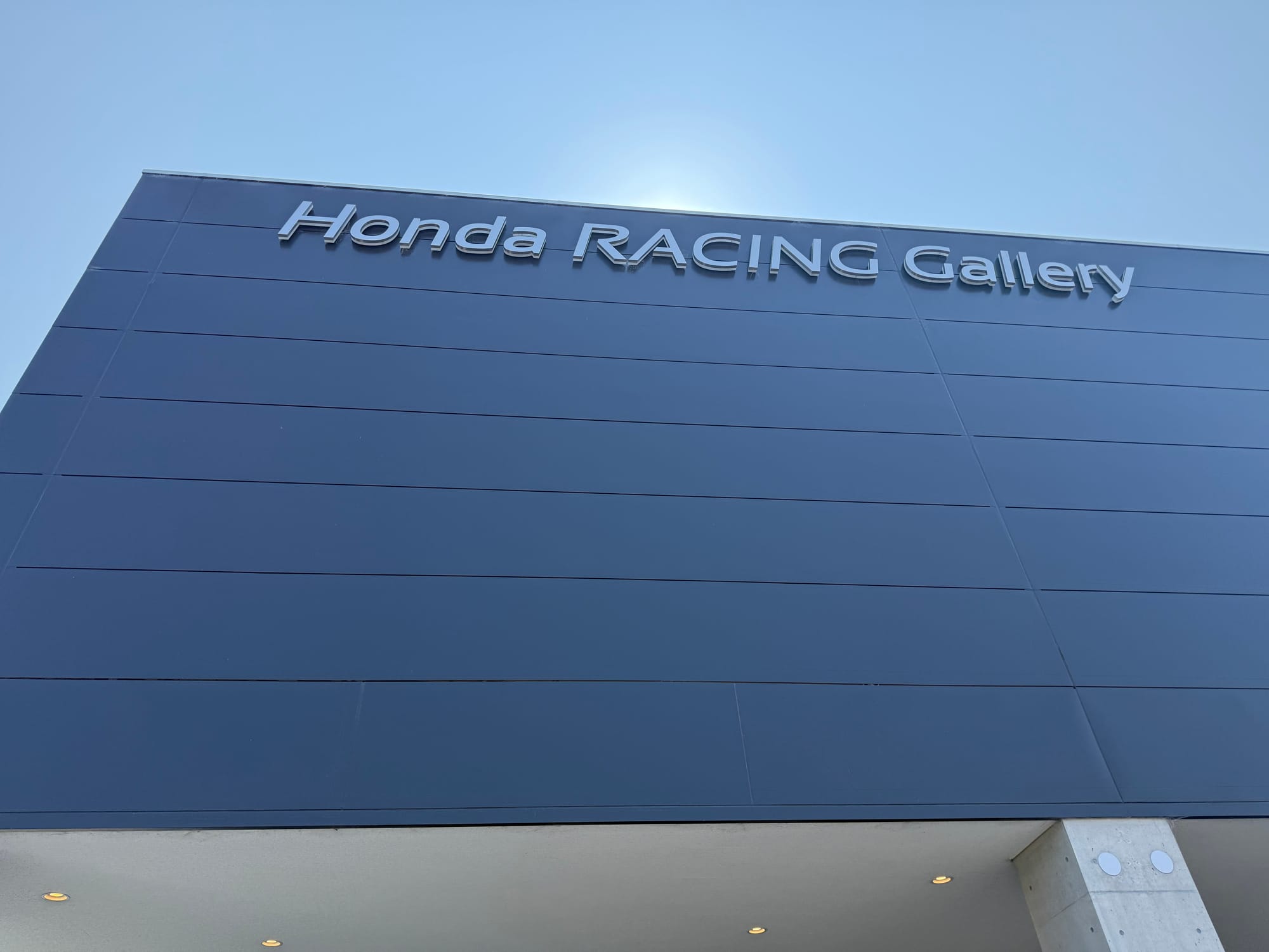 Exterior of the Honda Racing Gallery at Suzuka Circuit, featuring the museum's dark blue façade and silver lettering beneath a clear summer sky.