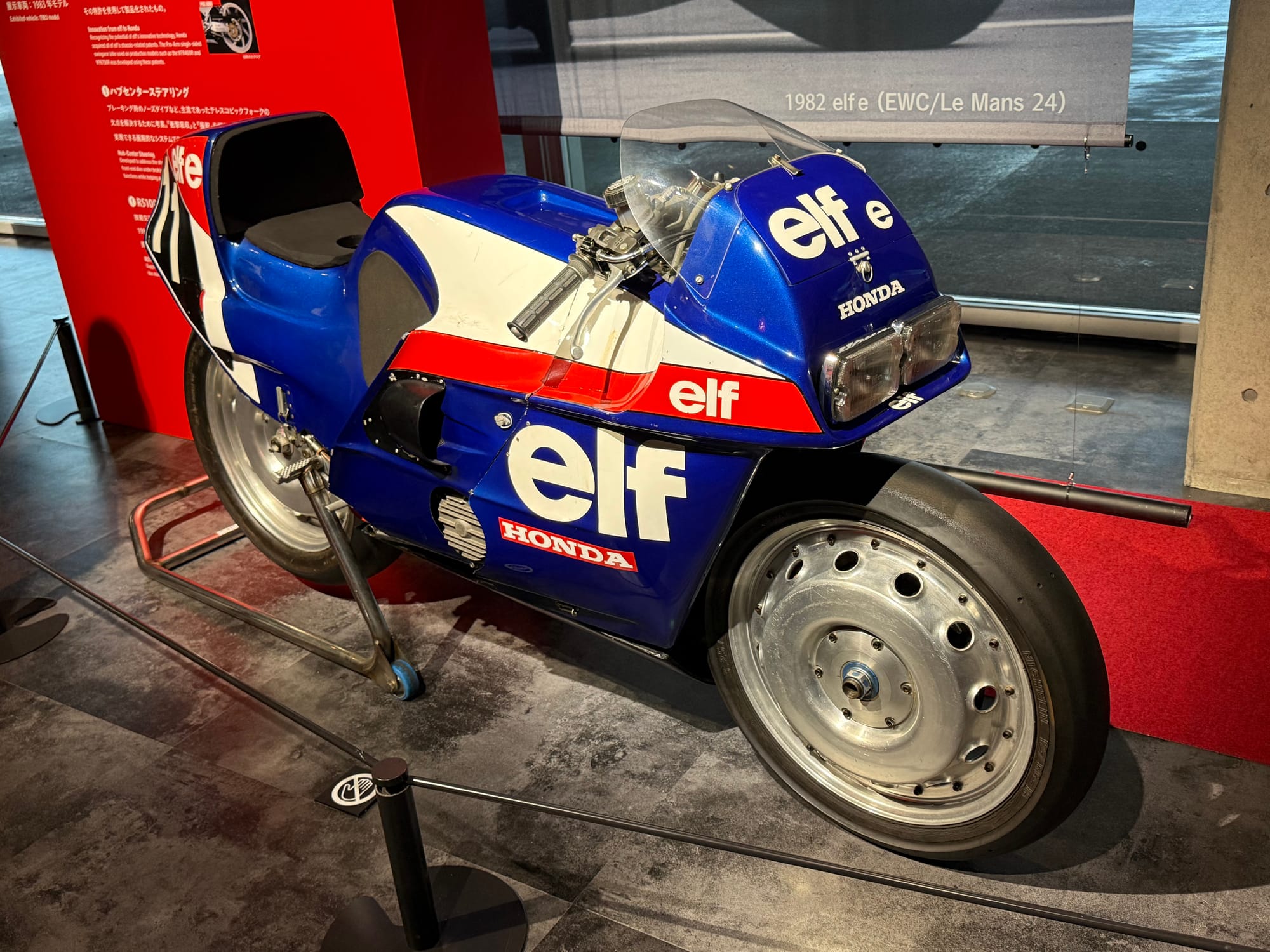 Honda elf endurance racing motorcycle displayed at the Honda Racing Gallery, featuring enclosed wheels, aerodynamic bodywork and the blue, white and red elf livery used in 1982.