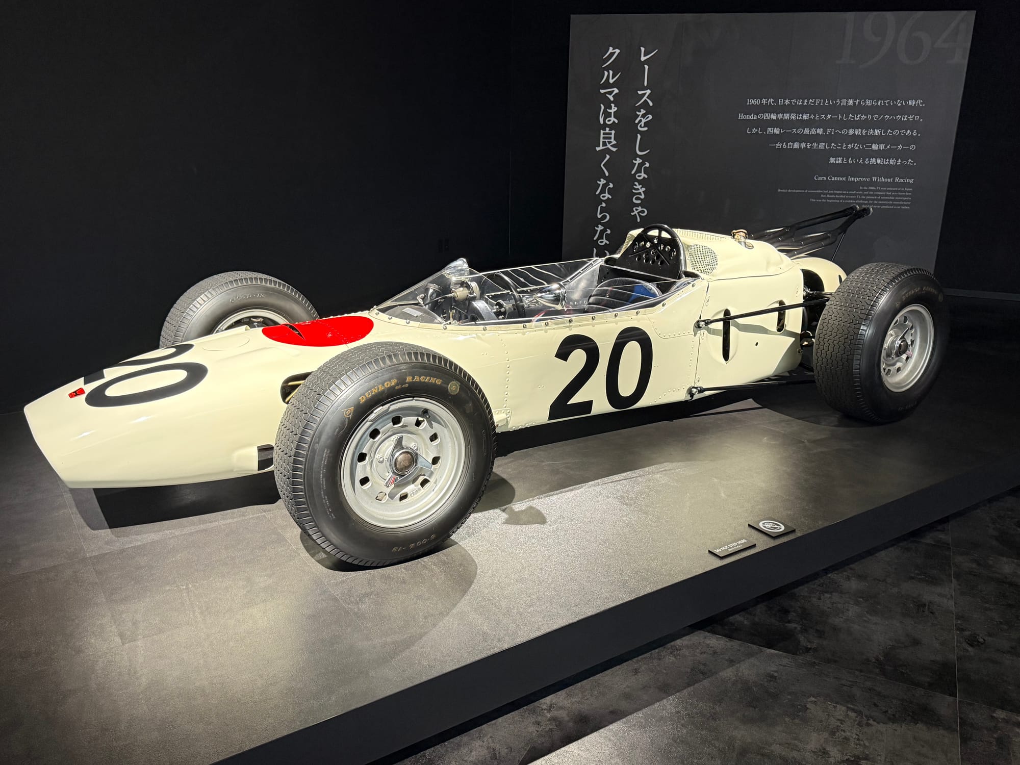 Honda RA271 Formula One car displayed at the Honda Racing Gallery, featuring white bodywork with a red sun motif and the number 20, Honda's first F1 machine from 1964.
