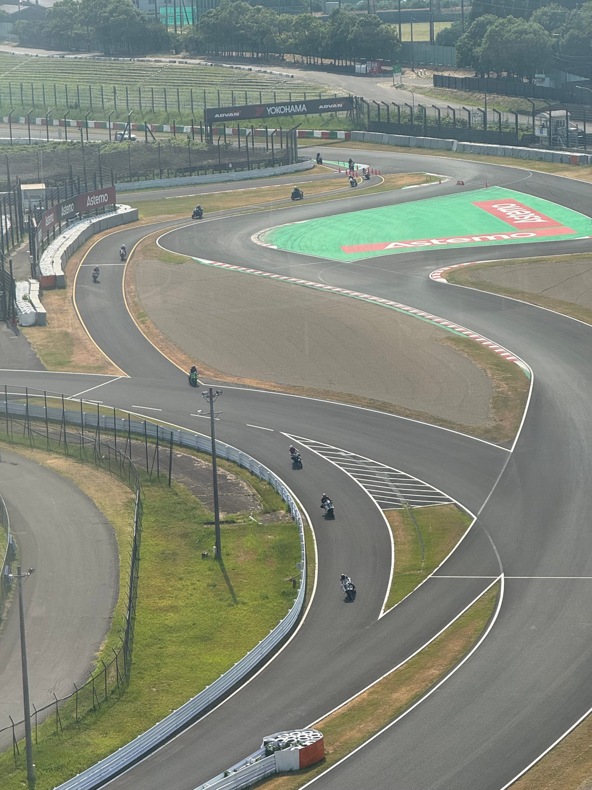 Motorcyclists ride through a section of the Suzuka Circuit during a two-wheel practice session, viewed from above as riders negotiate the circuit's sweeping corners.