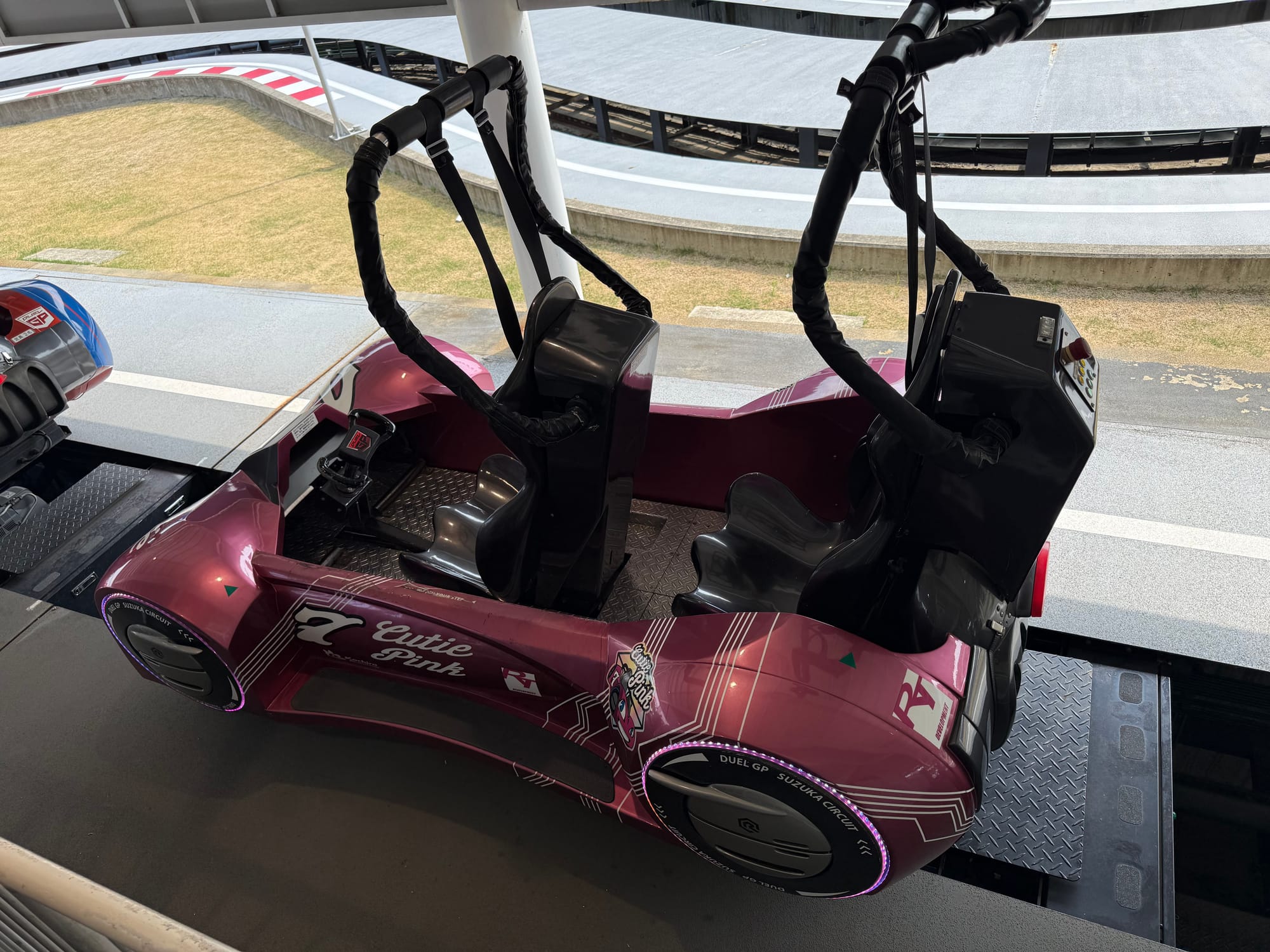 Pink Dual GP ride vehicle named Cutie Pink at Suzuka Circuit Park, featuring two seats, racing controls and a futuristic design beside the racing track.