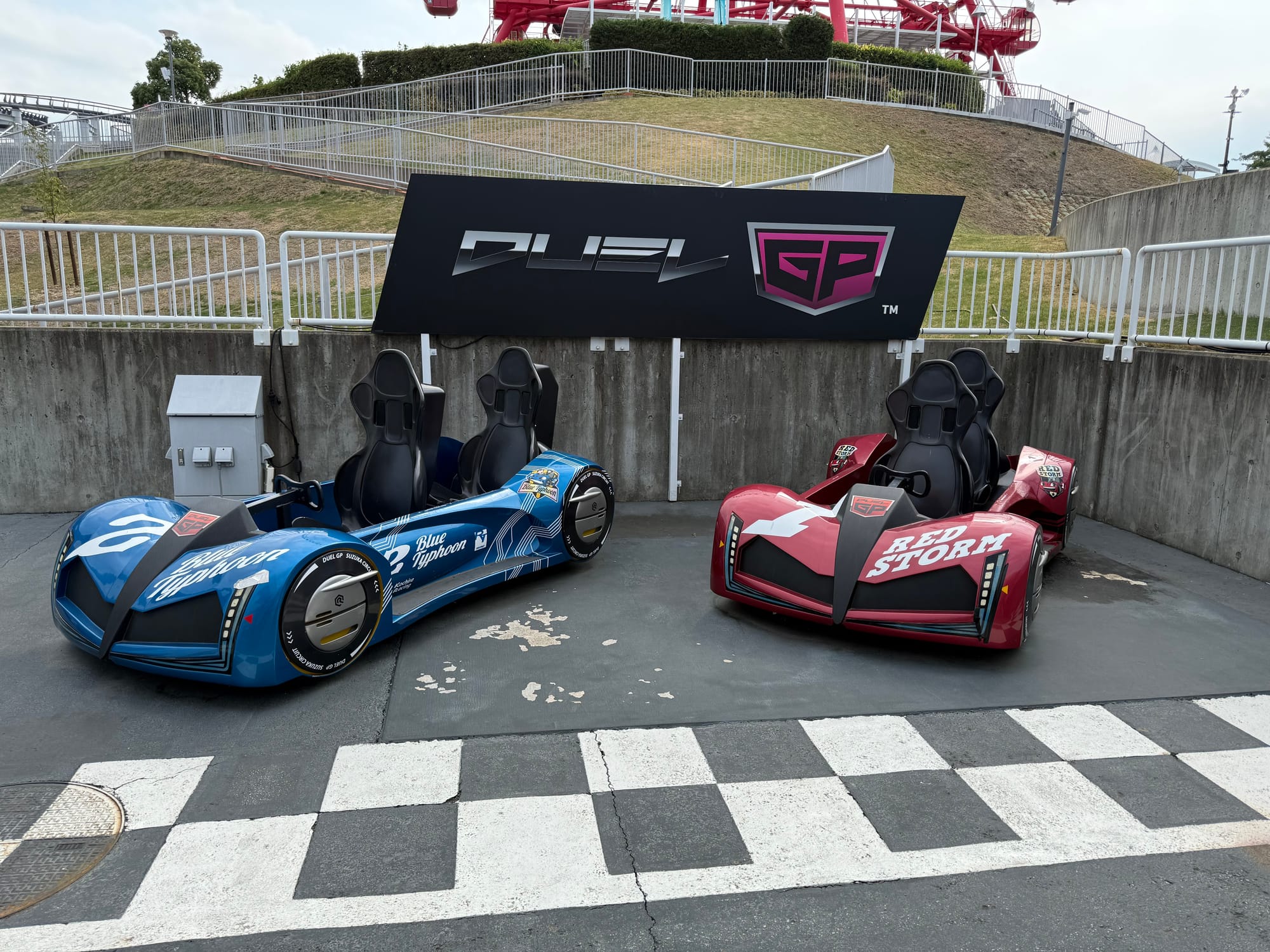 Dual GP ride vehicles at Suzuka Circuit Park, including the blue Blue Typhoon and red Red Storm, displayed beside a chequered track-themed area.