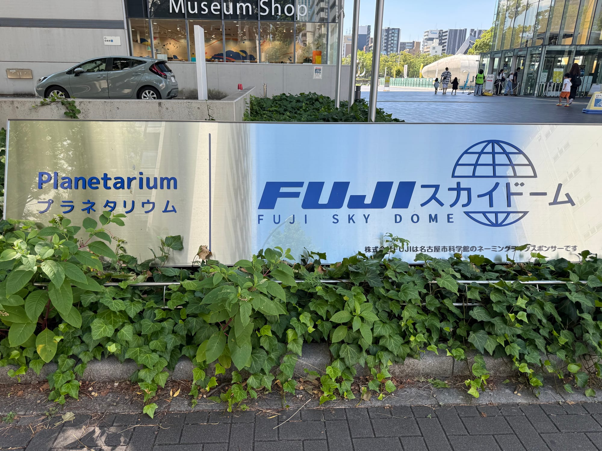 Copilot said:  Alt text (191 characters or under):  Sign for the Fuji Sky Dome planetarium at Nagoya City Science Museum, featuring English and Japanese text, surrounded by greenery outside the museum entrance.