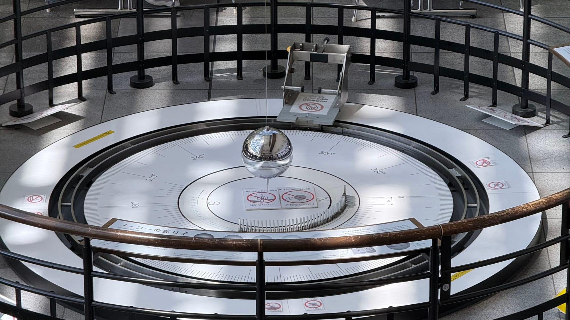 Foucault pendulum exhibit at Nagoya City Science Museum, featuring a suspended metal sphere swinging above a circular scale to demonstrate Earth's rotation.