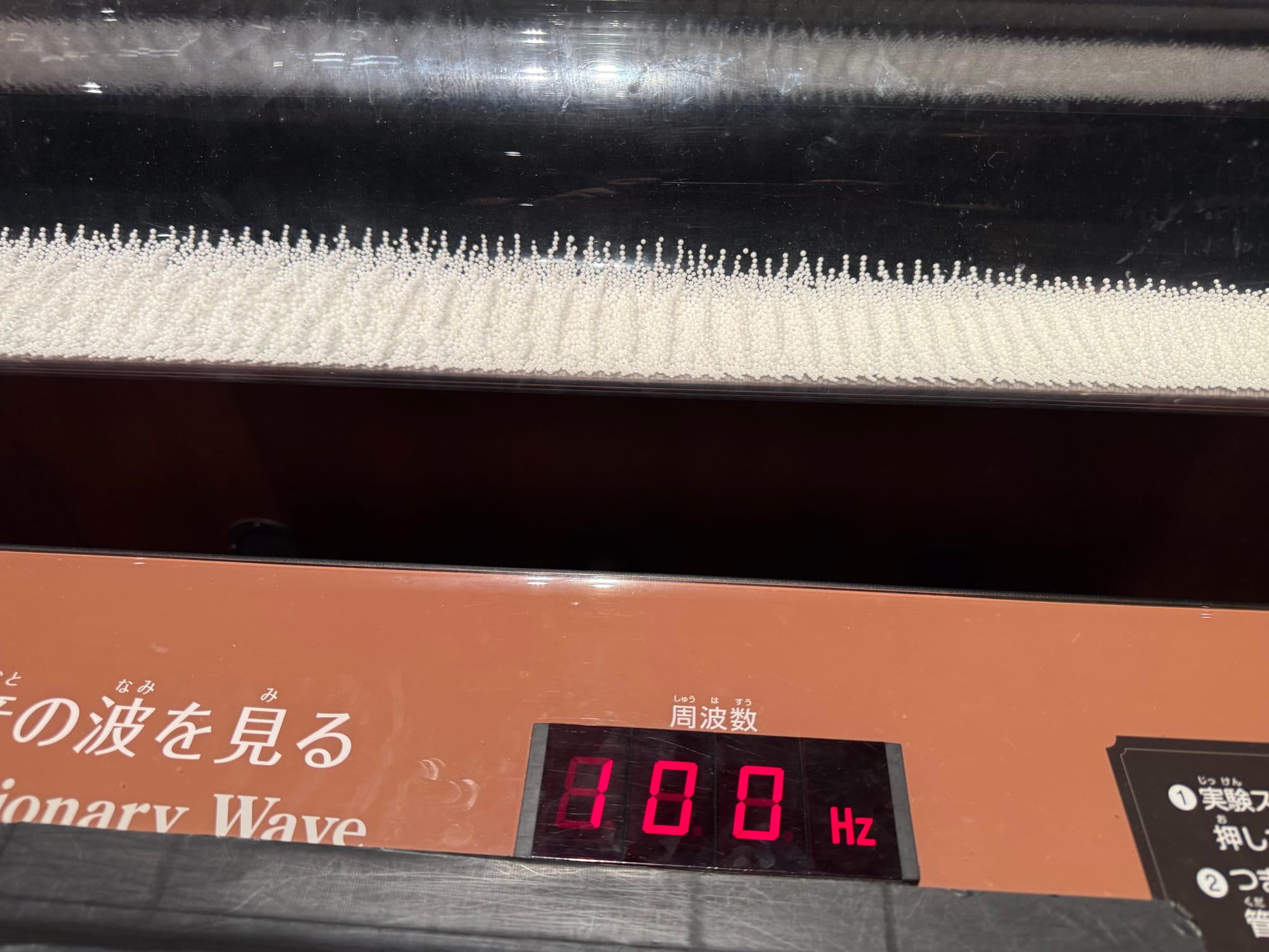 Standing wave exhibit at Nagoya City Science Museum, showing small white beads forming repeating patterns in a transparent chamber at a frequency of 100 hertz.