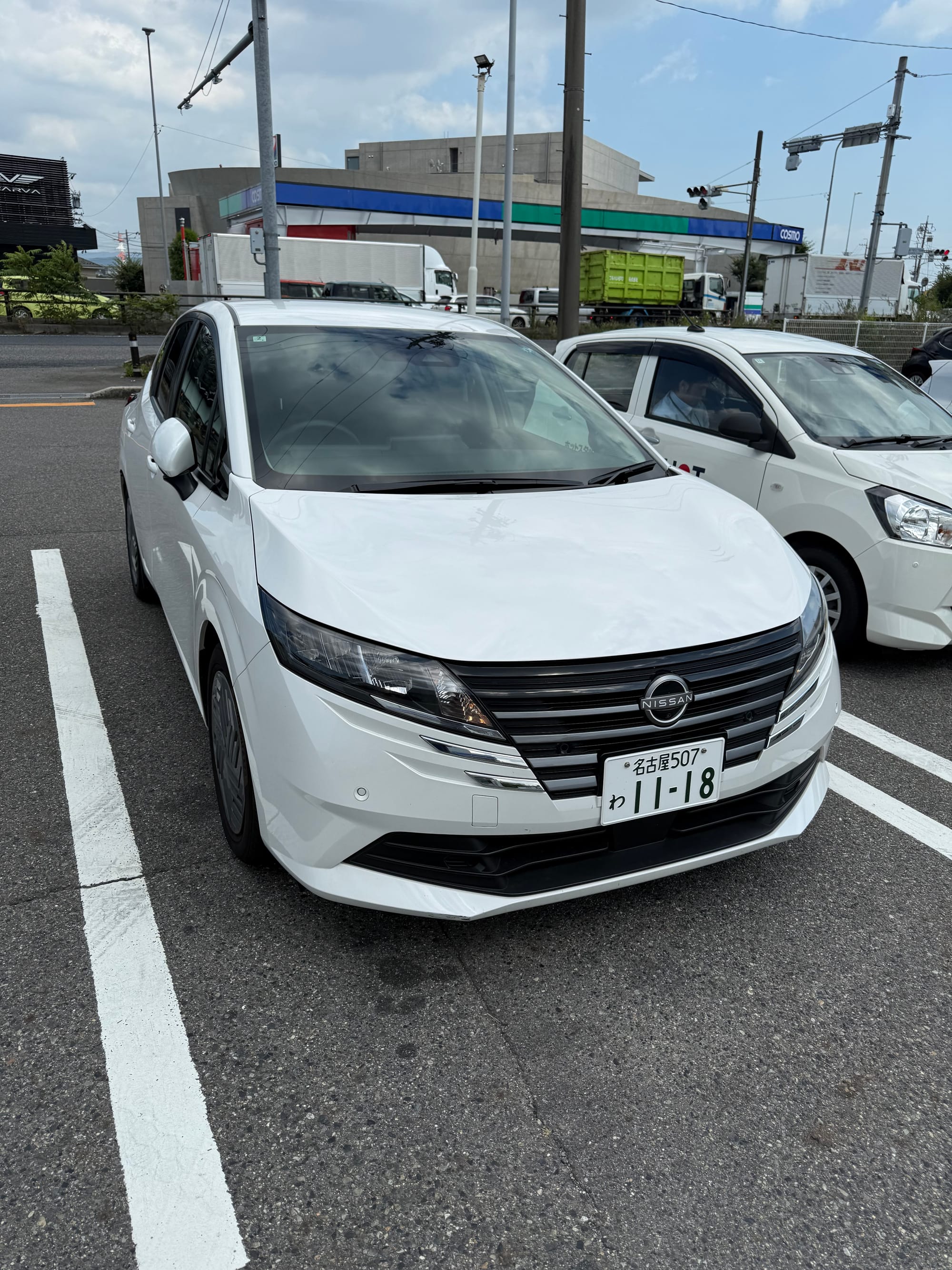 White Nissan Note e-POWER hatchback parked in a car park in Japan, viewed from the front, with a Japanese licence plate and roadside businesses in the background.