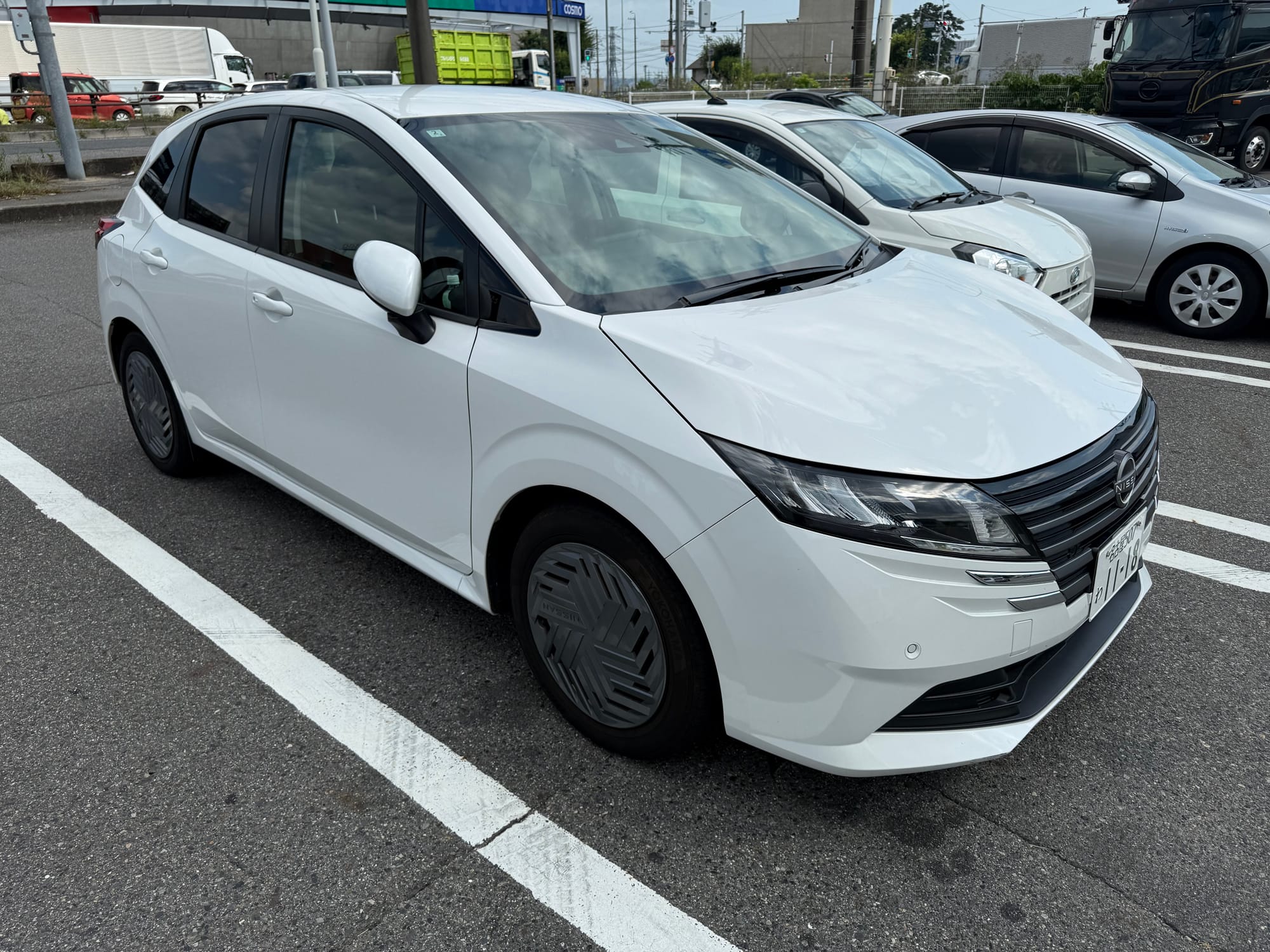White Nissan Note e-POWER hatchback parked in a car park in Japan, viewed from the front left, showing its aerodynamic bodywork and distinctive wheel covers.