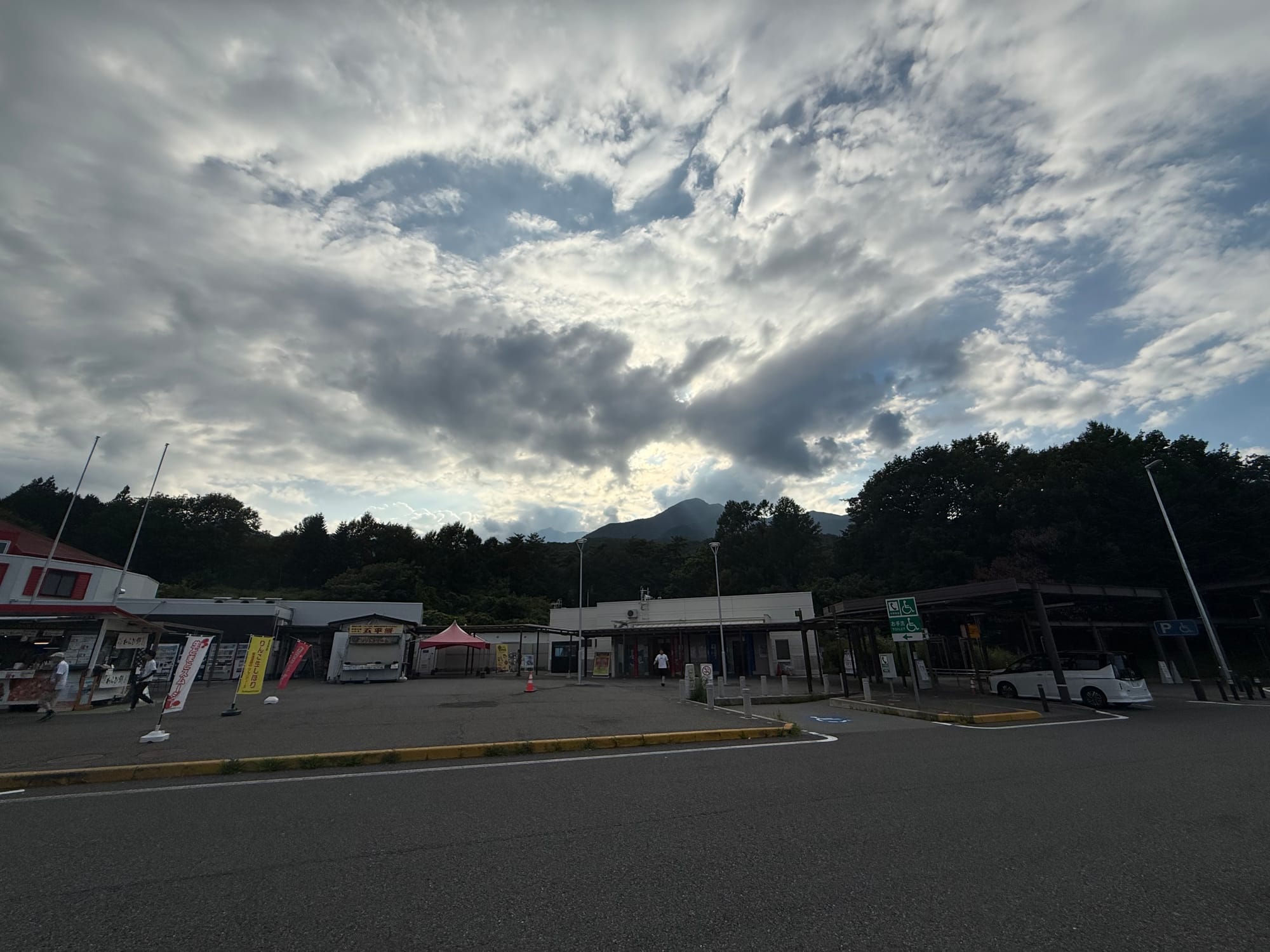 Mountain view from a Japanese expressway service area, with dramatic clouds gathering above forested hills and roadside facilities in the foreground.