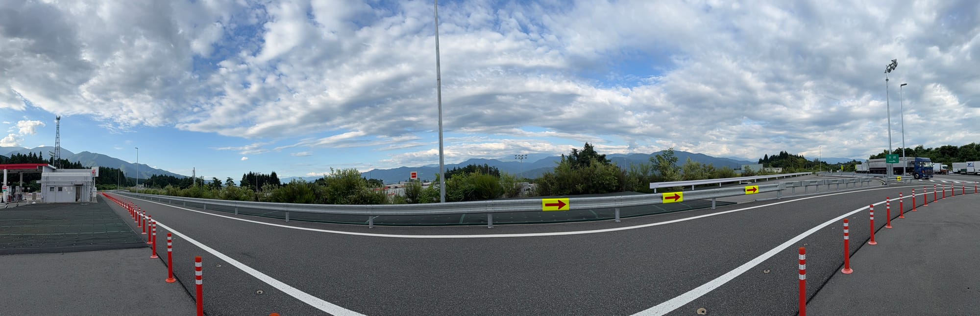 Panoramic view from a Japanese expressway service area, showing mountain ranges beneath dramatic layered clouds, with roads, trucks and roadside facilities in the foreground.